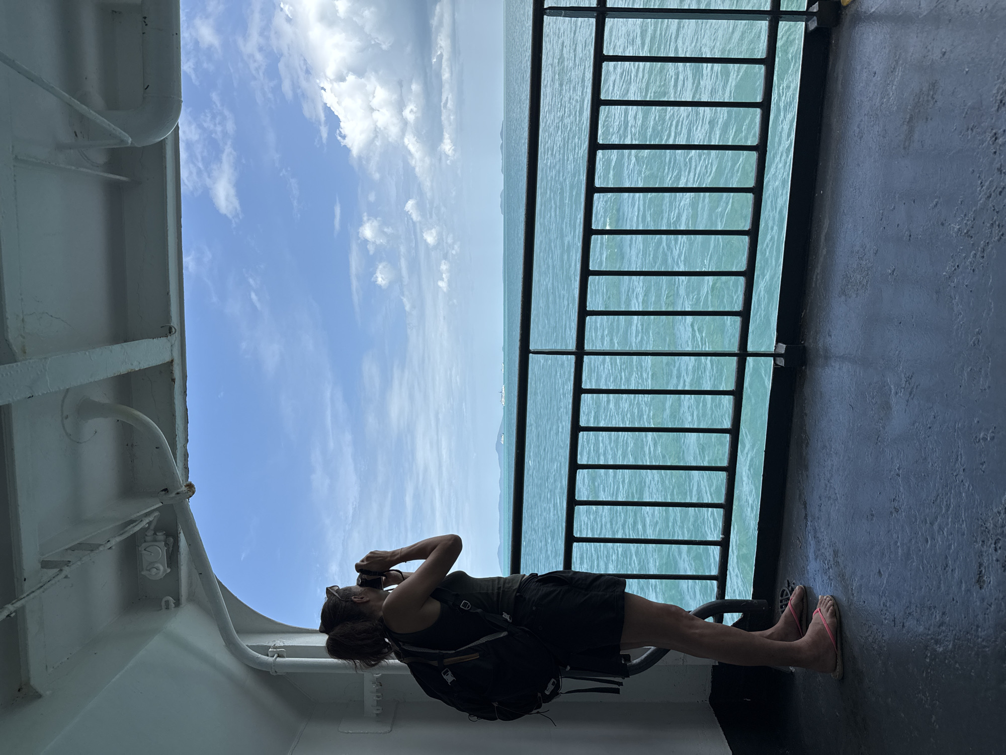 A woman with a ponytail and sunglasses is standing on a ferry, looking out at the ocean and holding a camera to her face. She is wearing a dark tank top, dark shorts, and pink flip-flops. A black backpack with orange accents is on her back. The ferry's deck is dark gray and wet. A black metal railing separates the deck from the turquoise ocean. In the distance, islands and several boats are visible under a blue sky with white clouds. The location is the Gulf of Thailand, near Don Sak, Thailand, as this is a common ferry route.