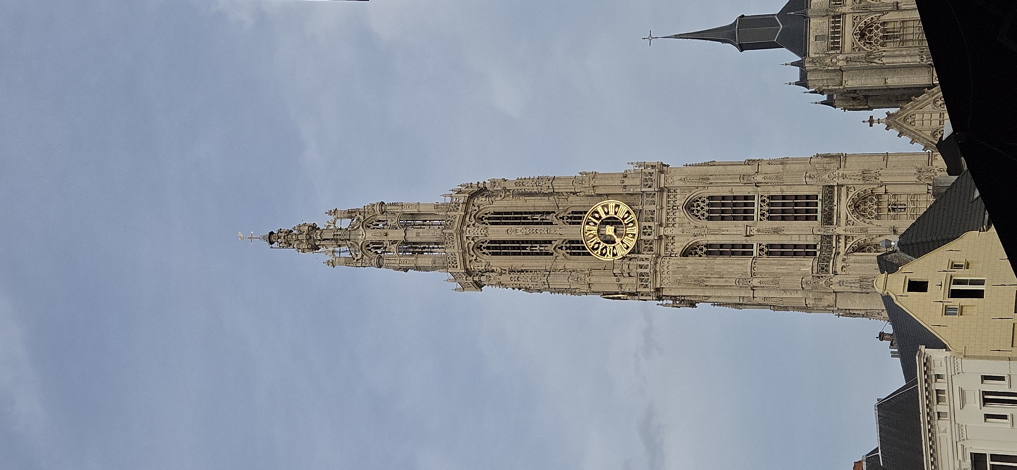 The image shows a detailed view of the tower of the Cathedral of Our Lady in Antwerp, Belgium, against a cloudy sky. The tower, constructed of light-colored stone, features intricate Gothic architectural details, including spires, arches, and decorative stonework. A large, ornate golden clock face with Roman numerals is prominently displayed on the tower. In the foreground and to the right of the tower, parts of other buildings are visible, including a smaller, darker spire with a cross. The scene is devoid of any people or discernible action. The overall environment is urban, with the focus being on the architecture of the cathedral.