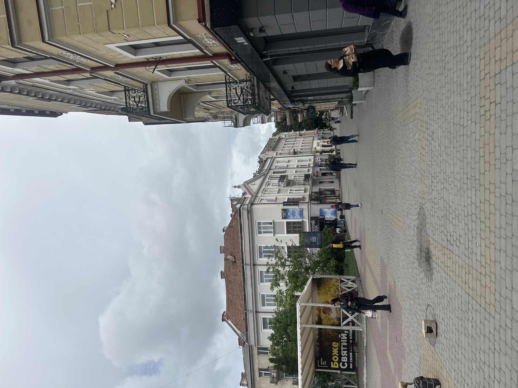 The image depicts a street scene in Ternopil, Ukraine, likely on a cloudy day. The viewpoint is from the street, looking down a cobblestone path lined with buildings on both sides. On the right, a yellow building with ornate balconies and decorative white trim dominates the foreground. On the left, a lighter colored building with a terracotta roof and multiple windows is visible, with a covered kiosk in front of it displaying a banner with Ukrainian text and sunflowers. Several people are walking along the street. In the foreground on the right, a woman in a black outfit is walking away from the camera, carrying a paper bag. Further down the street on the left, a woman in a black jacket and pants is walking towards the camera. Other individuals are scattered throughout the scene, walking in various directions. The environment appears to be a commercial or residential area with shopfronts visible on the ground floors of the buildings.