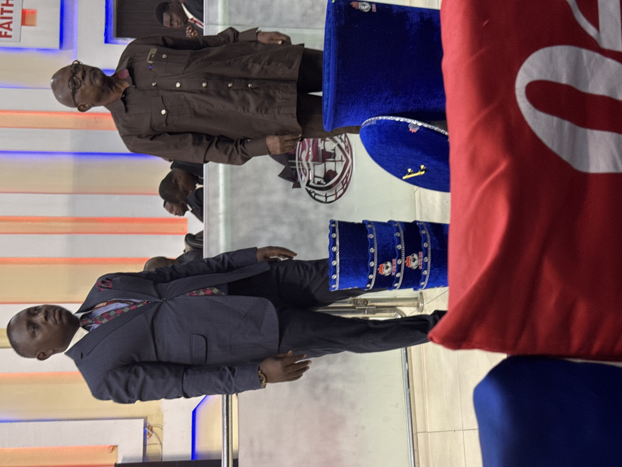 The image shows two men standing in what appears to be a building interior. The man on the right wears a dark suit and a patterned tie. The man on the left wears a brown collared shirt and eyeglasses. The scene includes two blue, tiered, ceremonial hats, a red object that is possibly a flag or large decorative pillow, and a logo displayed in the background. It is possible this photograph was taken in the city of Lugbe, Nigeria. There are multiple people present in the background.
