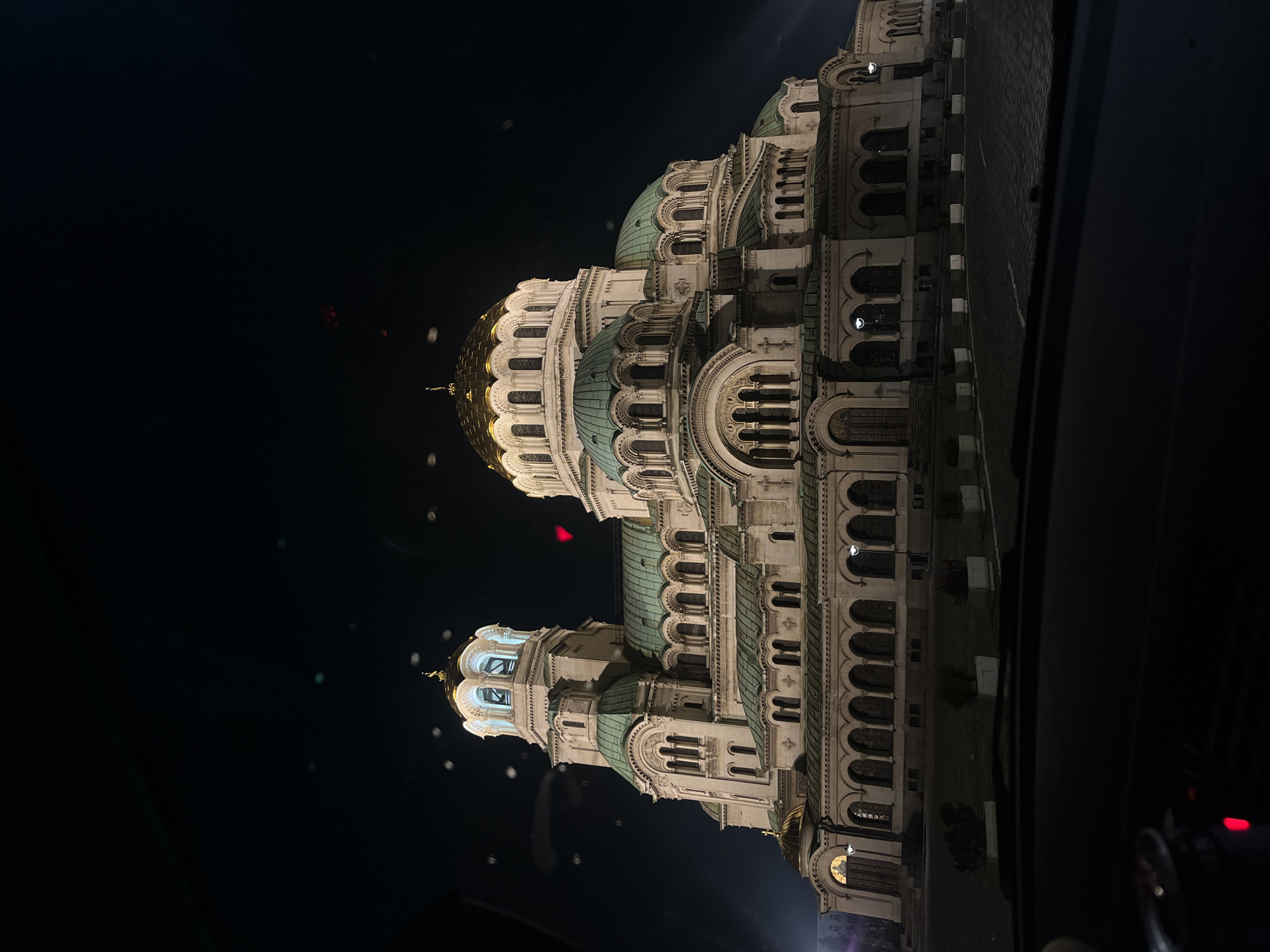 This media file captures the majestic St. Alexander Nevsky Cathedral in Sofia, Bulgaria, at night. The grand Eastern Orthodox cathedral is the primary subject, brilliantly illuminated against a deep black sky. The image is taken from a slightly low angle, seemingly from inside a vehicle, with parts of the car's interior visible as dark frames along the right and top edges.
The cathedral itself is a study in ornate architecture, characterized by its numerous domes and towers. Several large domes are topped with gleaming golden cupolas and crosses, while others feature distinctive green or turquoise-colored roofs. The building's facade is constructed from light-toned stone, richly detailed with arched windows, decorative columns, intricate moldings, and various architectural embellishments. Many of the cathedral's windows are lit from within, casting a warm glow that highlights the structural elements.
The time of day is clearly night, with the sky appearing uniformly dark, emphasizing the cathedral's radiant illumination. Scattered across the dark areas of the image are several small, circular light reflections or lens flares, possibly indicating light sources outside the camera's direct view or reflections off a window. A faint red light, likely a car's taillight, can be discerned in the lower right corner, reinforcing the impression that the photograph was taken from a moving or stationary vehicle. There is no discernible activity or event taking place, and no visible text. The overall atmosphere conveys a sense of awe and reverence for the impressive architectural landmark.