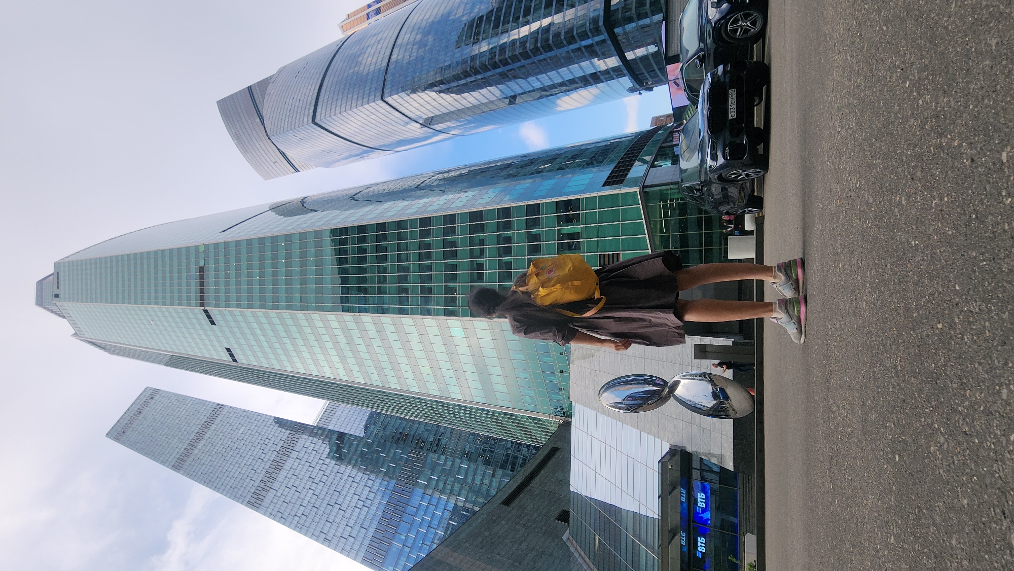 A medium-shot, rotated image shows a person, a yellow backpack on their back, standing near a sidewalk next to several high-rise glass office buildings. The buildings are in Moscow, Russia, and are part of the Moscow International Business Center, also known as Moscow City. The person is wearing a dark-colored dress and tennis shoes. Cars are parked on the side of the road. Two large, reflective, teardrop-shaped sculptures stand between the person and the buildings.