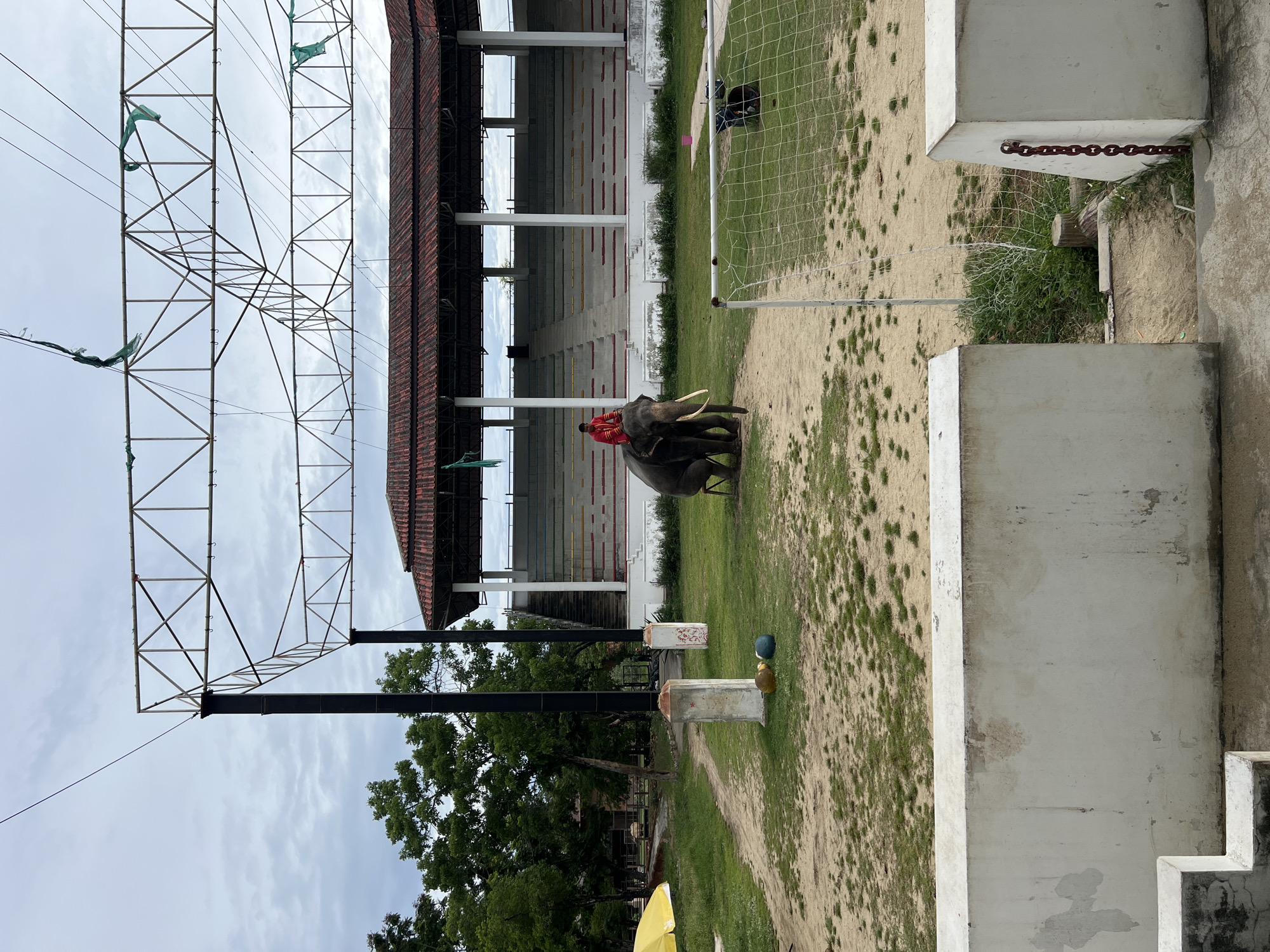 The image depicts an outdoor scene in Bang Pu Subdistrict Municipality, Thailand, likely at a circus or performance venue. In the foreground, a white concrete structure with visible wear and tear forms a partial barrier. A dirt path with patches of green grass leads towards the center of the image, where a large elephant stands. A person in a red top is seated on the elephant's back. Behind the elephant is a grandstand with multiple rows of seats, some of which are colored red, yellow, and green, suggesting it's a seating area for spectators. Above the grandstand, a metal truss structure with wires and torn green netting is visible against a cloudy sky. To the right, a white net, possibly part of a sports field, extends into the frame. The overall environment appears to be a somewhat weathered outdoor entertainment or sporting facility.
