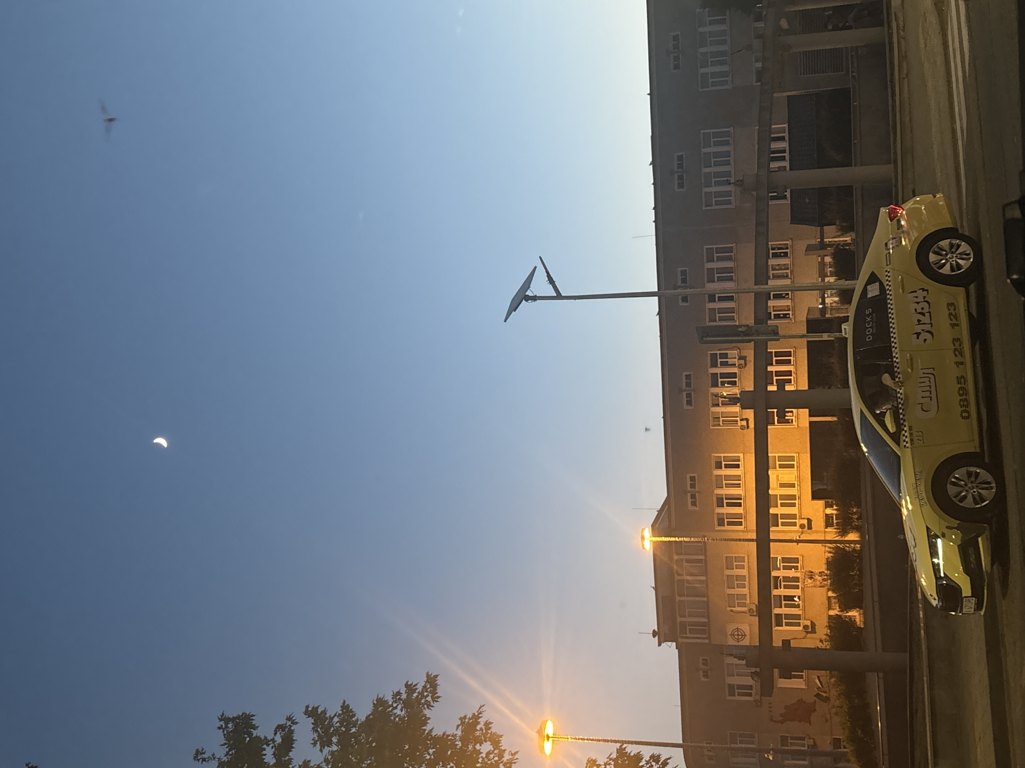In Burgas, Bulgaria, a yellow taxi is visible driving on a road in front of a building during twilight. The sky is clear and blue, with a crescent moon visible in the upper left quadrant and a flying insect in the upper right. Two streetlights with glowing orange lights are positioned in the foreground. The building behind the taxi is multi-story with numerous windows. The taxi has the number "S1234" displayed prominently on its side, along with a phone number and the word "DOCKS". The taxi's headlights and taillights are illuminated.