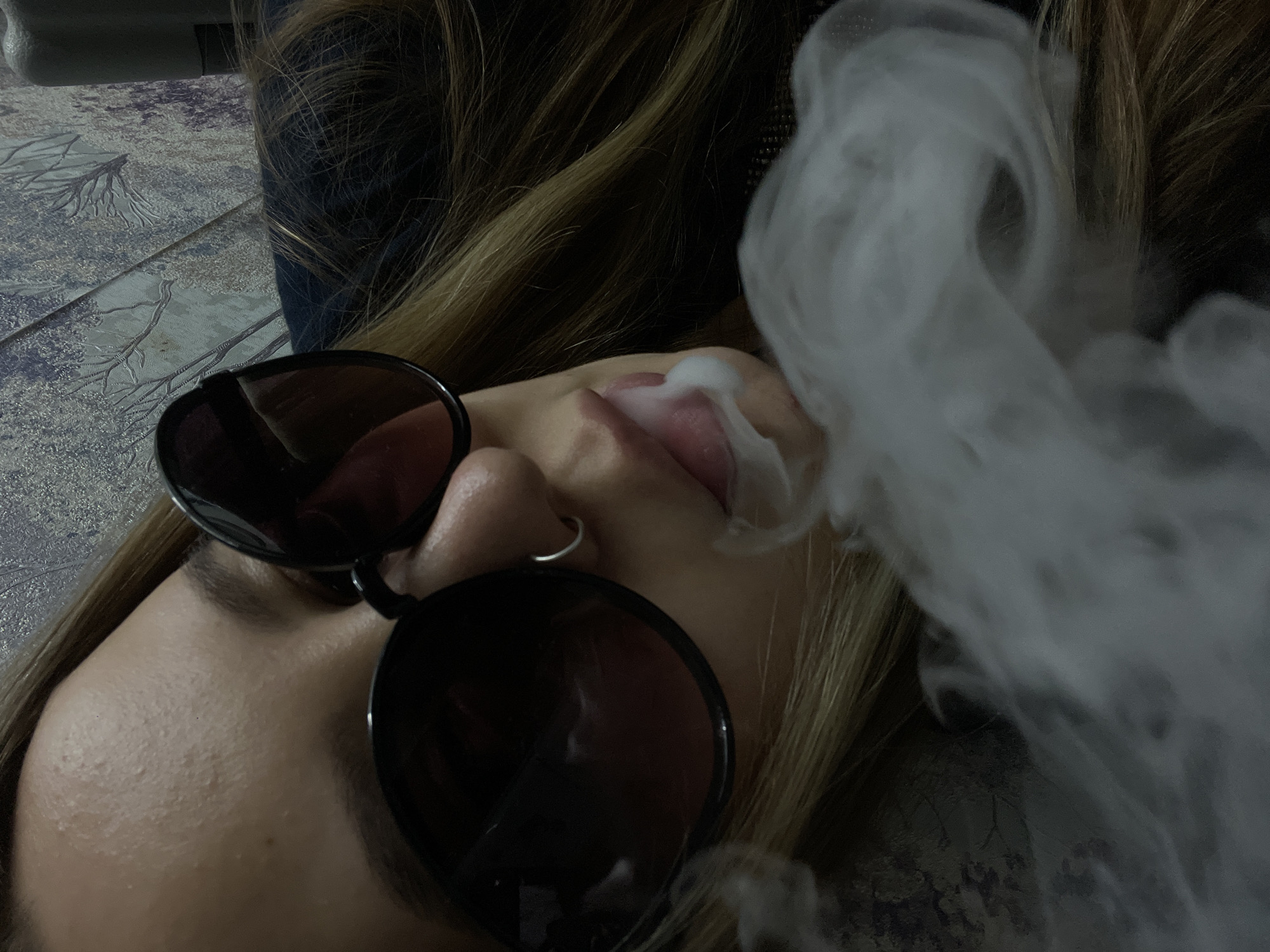 A close-up, angled shot of a person with sunglasses exhaling smoke. The person has light brown hair, fair skin, and a nose ring. The sunglasses are round and dark, obscuring the eyes. The smoke is white and billowy, partially obscuring the face. The background is blurred and indistinct, but appears to be a patterned fabric or surface. There is no information to suggest this photo was taken in Novopokrovskaya, Russia.