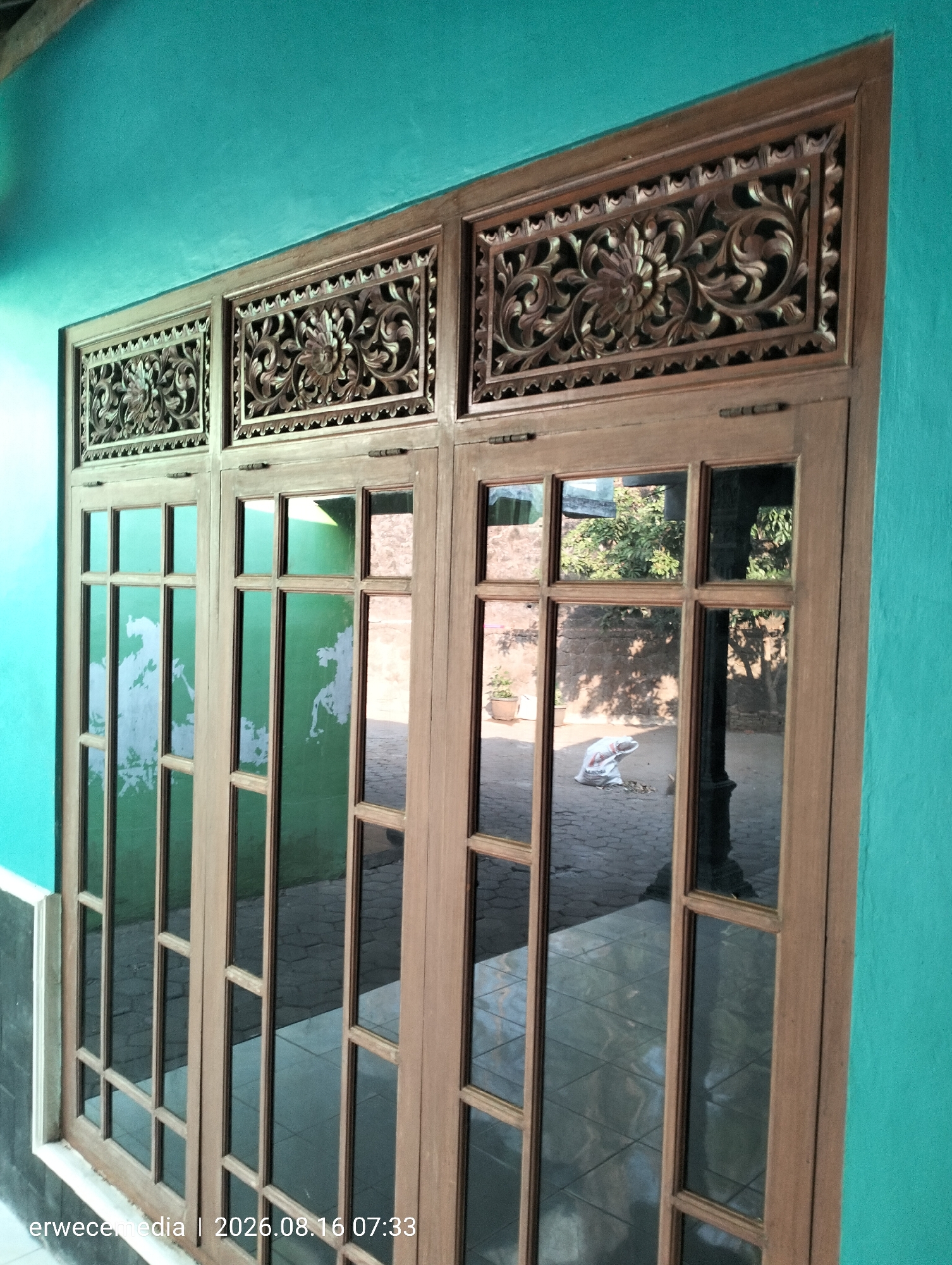 The image shows a close-up view of a wooden-framed window with carved wooden panels above the glass panes. The window is set against a teal-colored wall. The window has multiple glass panes, and the reflections in the glass show an outdoor scene with trees, a brick-paved ground, and a white bag. The lighting suggests it is daytime. The timestamp "erwecemedia | 2026.08.16 07:33" is visible in the bottom left corner of the image, indicating the time and source of the photograph. The location is specified as Imogiri, Indonesia.