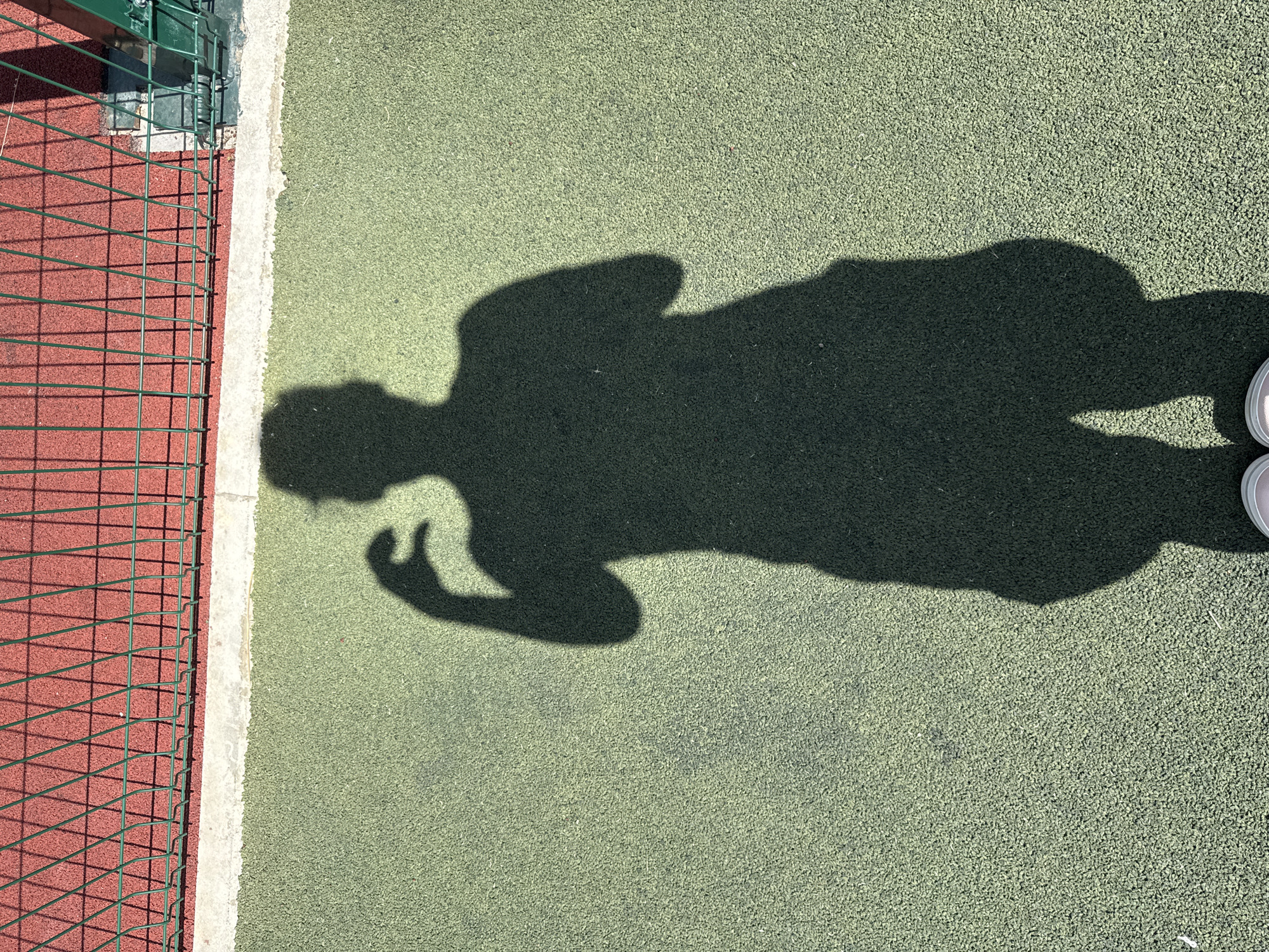 The image shows a shadow of a person on a green, textured outdoor surface, likely a sports court. The shadow depicts the person from the waist down, with one arm bent at the elbow and the hand raised as if to take a photo or make a gesture. The person appears to be wearing shorts. In the background, there is a red surface behind a green metal fence, casting shadows on the red surface. The scene is outdoors under sunlight, indicated by the distinct shadows. The location in the city of Vyazniki, Russia, is not explicitly depicted in the image itself, but is provided as contextual information.