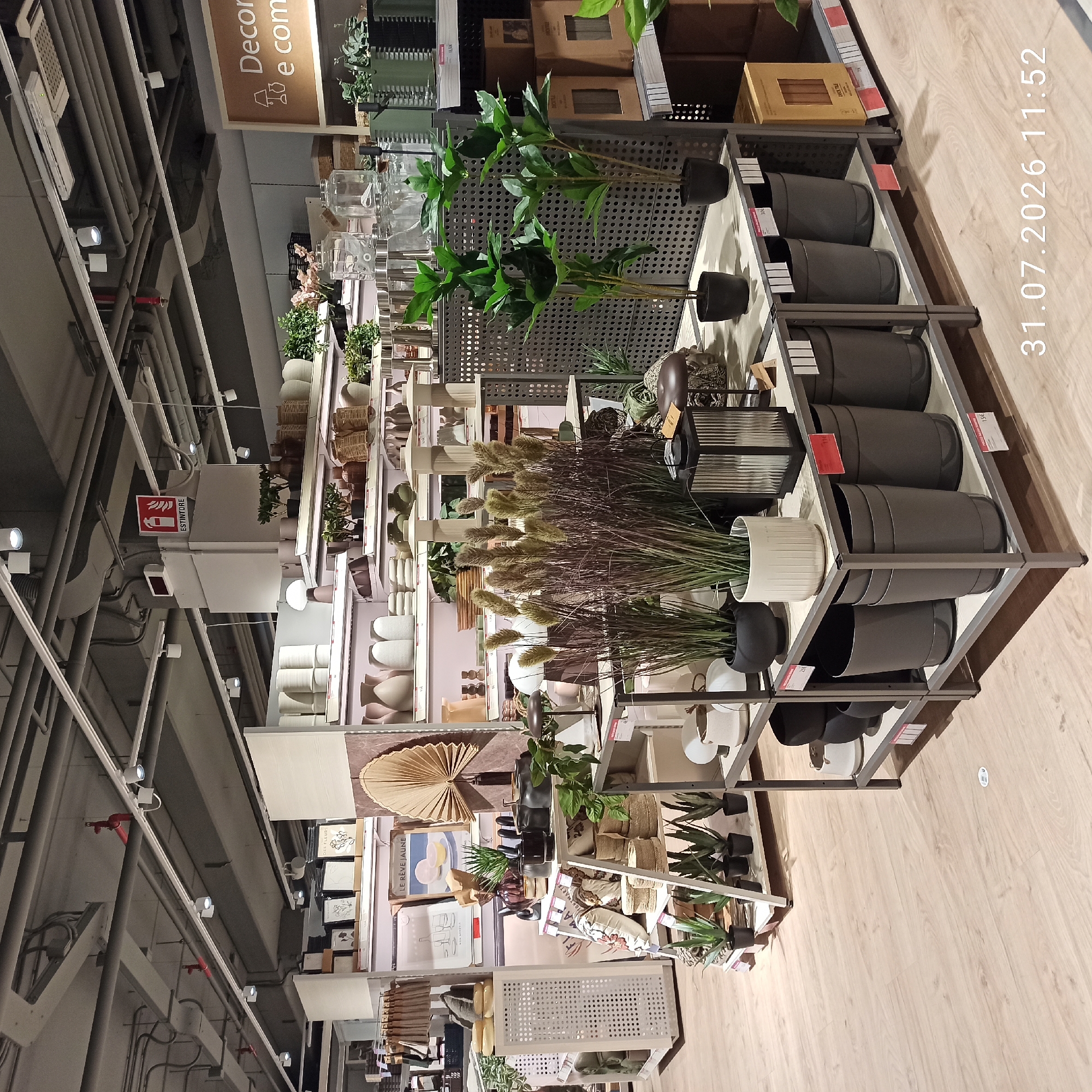 The image shows an indoor retail environment, likely a home decor or furniture store, with a focus on plants and decorative items. The scene is well-lit, with overhead track lighting illuminating the display shelves. On the shelves, various items are arranged, including potted plants, vases, baskets, lanterns, and decorative pillows.
In the foreground, a shelving unit displays several grey plant pots and a white ceramic planter with ribbed detailing. A black metal lantern with glass panels sits on the top shelf of this unit. To the right of this display, a shelving unit holds more decorative items, including potted plants and a box labeled "DECK TILE".
In the mid-ground, a larger display unit features tall, feathery grasses in large planters. Behind this, more shelves are stocked with an assortment of ceramic vases in neutral tones. To the left of the image, white storage units and shelves display more decorative items, including framed art prints and pillows.
In the background, the ceiling structure is visible, with exposed pipes and conduits. A red fire extinguisher sign with the text "ESTINTore" is mounted on a white pillar in the upper mid-ground. Further back, more shelves are visible, stocked with various home goods.
A timestamp in the lower right corner indicates "31.07.2026 11:52".
Based on the retail context and the style of the items displayed, this scene is consistent with a store like IKEA. Given the presence of the timestamp and its proximity to the date "2026", this image was likely taken in one of IKEA's locations. Without additional external information, pinpointing the exact city of Modugno, Italy, is not possible solely from the visual content of the image. However, if this image were to be associated with Modugno, it would be within a commercial establishment within that municipality.