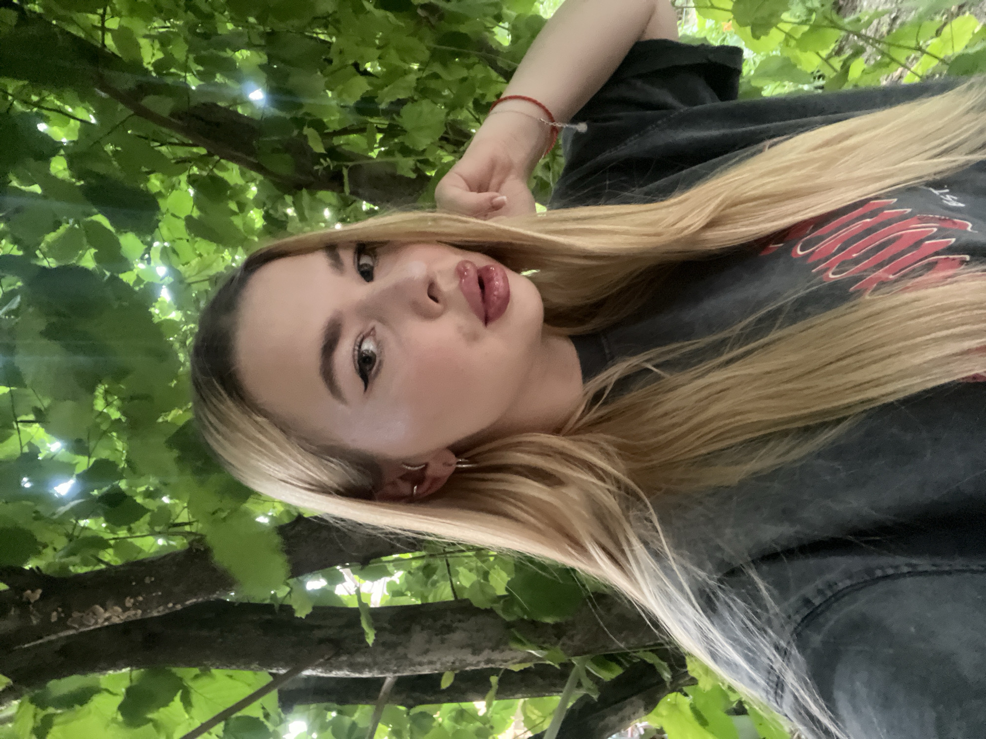 A young woman with long blonde hair and dramatic eyeliner poses for a selfie outdoors, surrounded by trees and green foliage. She is wearing a dark, distressed t-shirt with red lettering. Her left hand is resting on her cheek, and she has a red string bracelet on her right wrist. The background suggests a natural setting, possibly a park or a wooded area. There are no discernible landmarks indicating the city of Tikhoretsk, Russia.