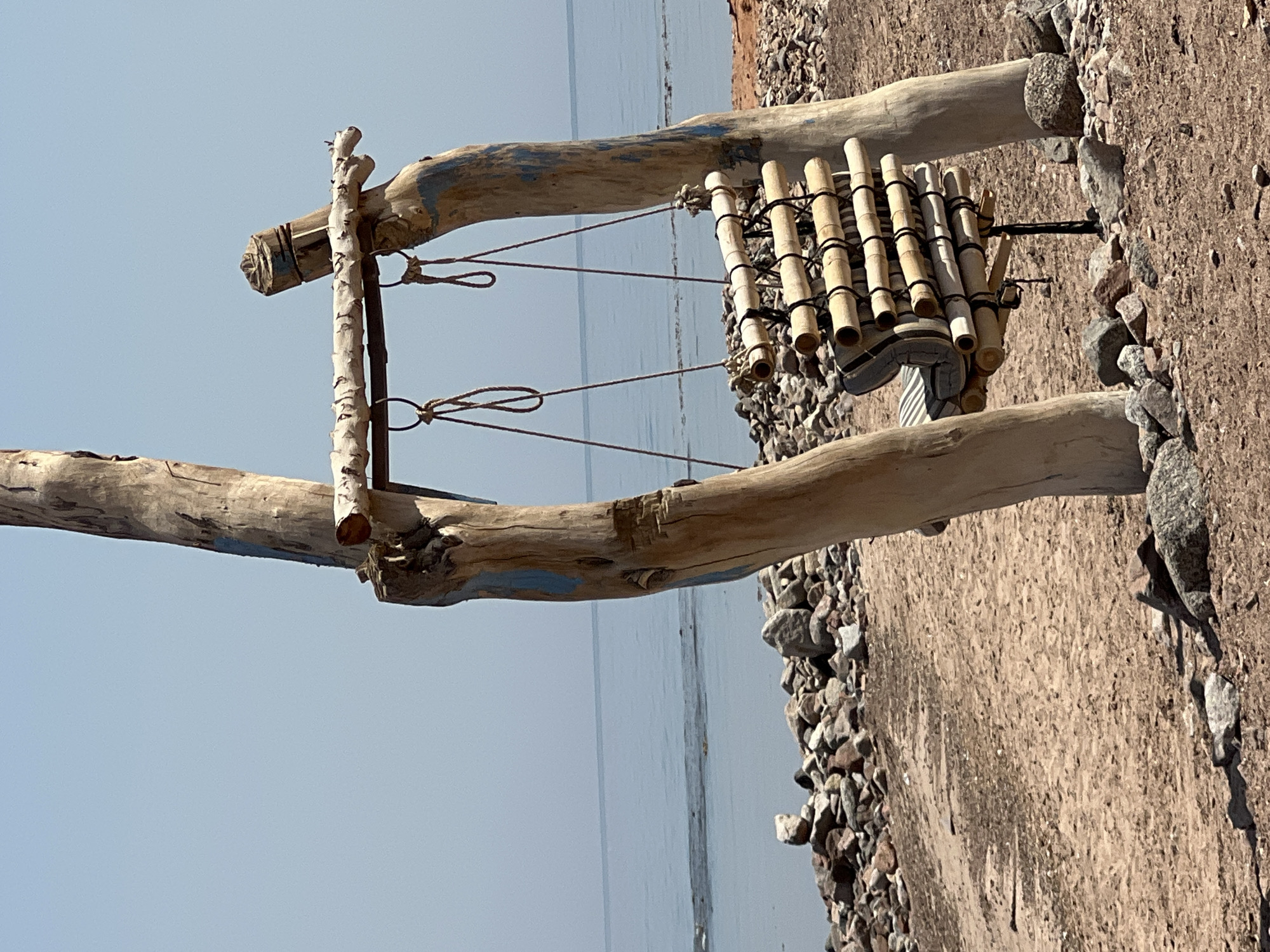 The image depicts a rustic swing constructed from driftwood, suspended between two upright wooden posts. The swing's seat is fashioned from bamboo poles secured with black bindings, and it appears to have a striped cushion. The swing is set against a backdrop of the sea and a rocky shoreline. The overall aesthetic suggests a natural, possibly remote, beach location. The sky is clear and a pale blue, meeting the calm, light blue water at the horizon. The ground is sandy and strewn with various sizes of rocks and stones, some of which are quite large. The driftwood posts supporting the swing have some blue paint markings on them. This scene is located in the city of Nueiba, Egypt. There are no people or animals visible in the image.