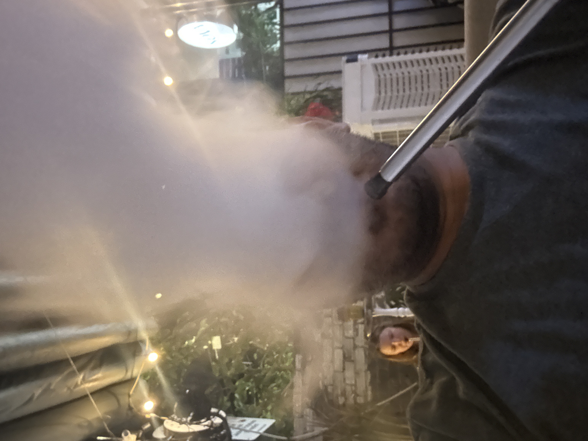 This rotated image captures a close-up of a person exhaling a voluminous cloud of white vapor or smoke, likely from a hookah or vape, in an outdoor setting during the evening in Đà Nẵng, Vietnam.
The main subject, partially visible on the left side of the frame (when oriented correctly), is wearing a dark grey shirt. A long, slender, metallic silver hose with a dark tip is held to their mouth, from which the dense cloud of smoke billows out, obscuring part of their head and drifting towards the left.
The background is softly blurred but reveals elements of a social outdoor environment. Warm yellow string lights are strung low across the scene, providing ambient illumination. Green foliage and what appears to be a brick wall are visible behind the smoke. To the right of the foreground subject, a white, slatted structure, possibly a fence panel or part of an outdoor unit, can be seen. Another person, with light brown hair, is partially visible in the lower right background, looking towards the activity.
At the top center-right, a circular, illuminated sign with indistinct light-colored text (possibly a stylized word like "OXYGN" or a brand name) suggests the location is a commercial establishment such as a cafe, restaurant, or lounge. The artificial lighting and general ambiance indicate it is nighttime or late evening. The scene conveys a relaxed and social atmosphere.