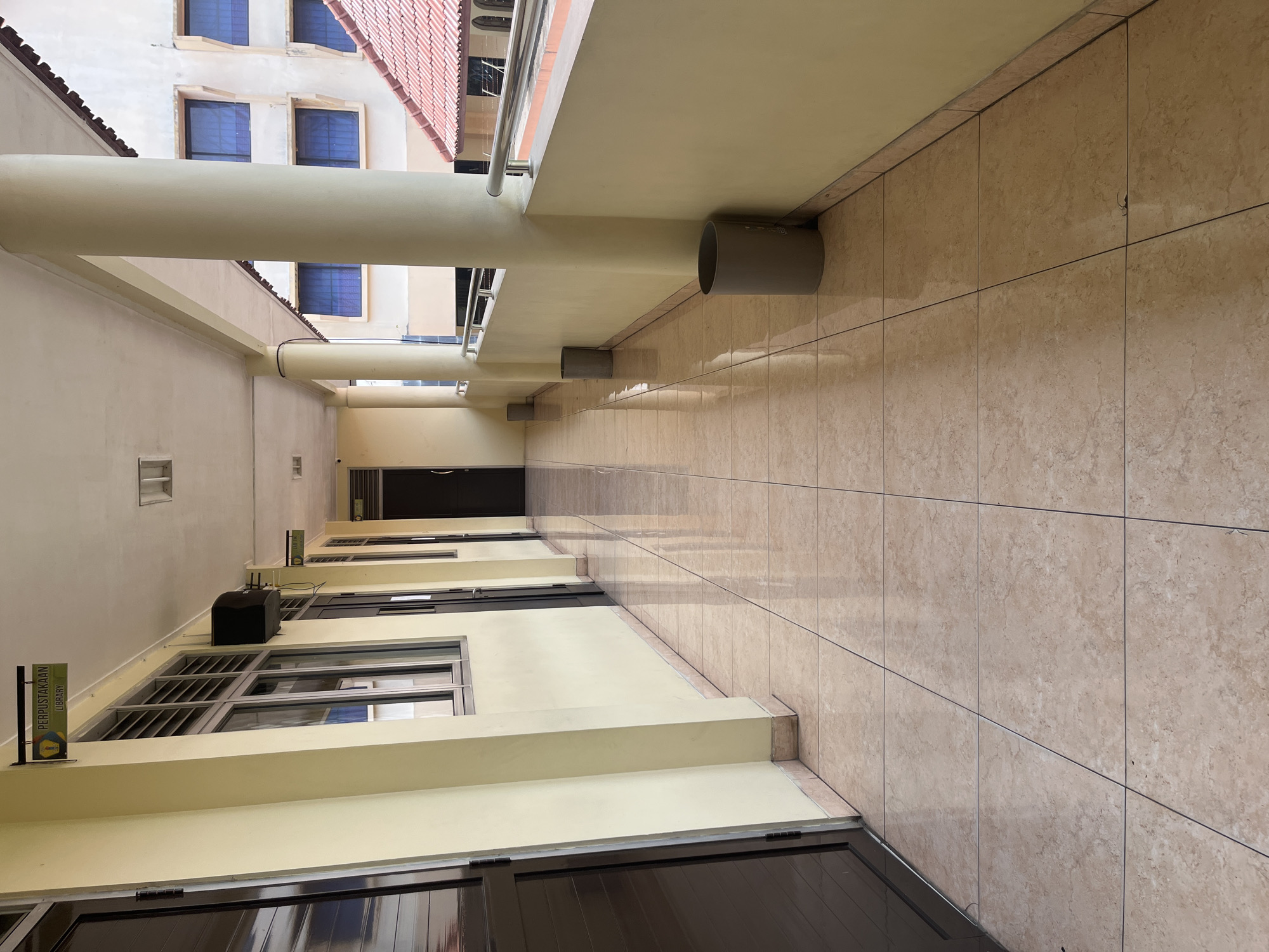 The image shows a sunlit, outdoor corridor with a tiled floor and a yellow wall on the left, which has several dark-colored doors. On the right side of the corridor, there is a railing and a tan-colored building with blue-tinted windows and a red tiled roof. The corridor is lined with several tall, cylindrical, gray trash cans. In the distance, a sign is visible hanging from the ceiling, indicating "PERPUSTAKAAN LIBRARY". The scene appears to be in a public building or institution, possibly a school or university, located in South Jakarta, Indonesia.