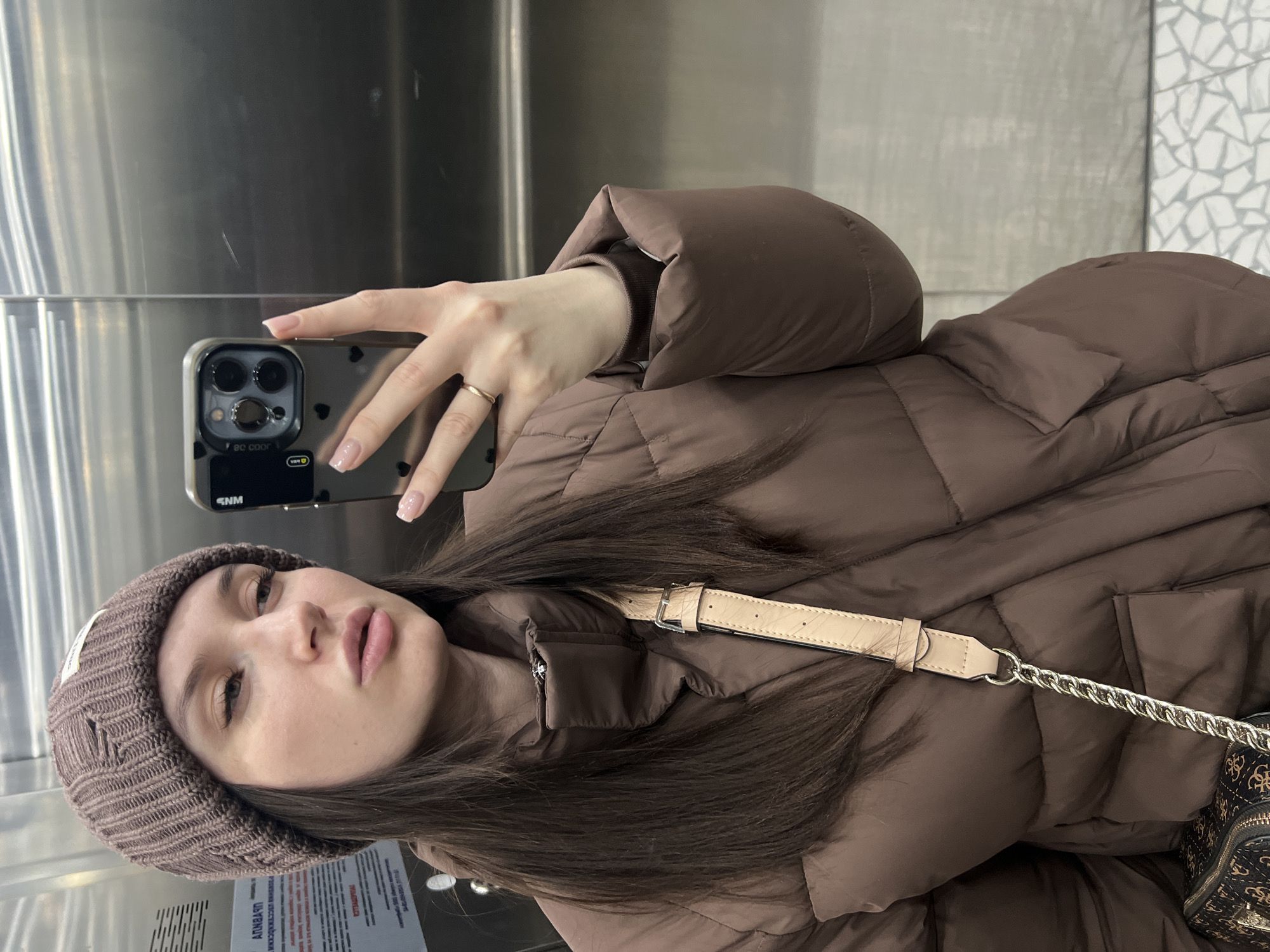 A woman takes a selfie in an elevator. She is wearing a brown puffer jacket and a brown knit hat. She holds an iPhone in her right hand. The background includes a stainless steel elevator interior and a wall with a mosaic pattern. The location is in Tyumen, Russia, based on visible text in Russian in the lower left corner.