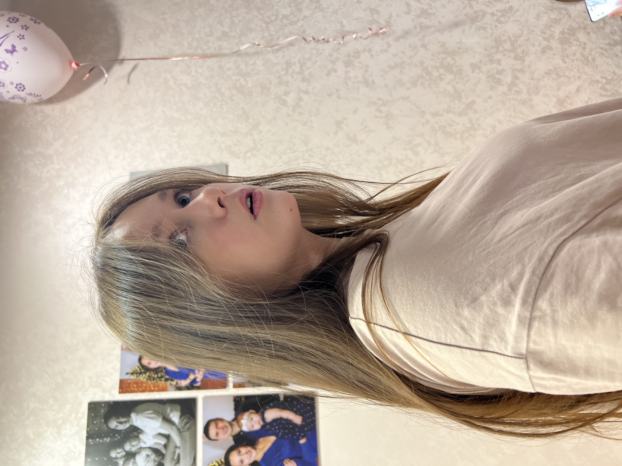 A close-up indoor shot depicts a light-skinned young person with long, light-brown hair, gazing upwards with slightly parted lips. The person is wearing a light-colored, long-sleeved garment. Above the person's head is a pink balloon with a butterfly design, and a curly ribbon. In the background, several framed photographs are visible on a wall with a textured, light-colored surface. Based on the family photos and other information, the location is likely in a residence in Chaikovsky, Russia.