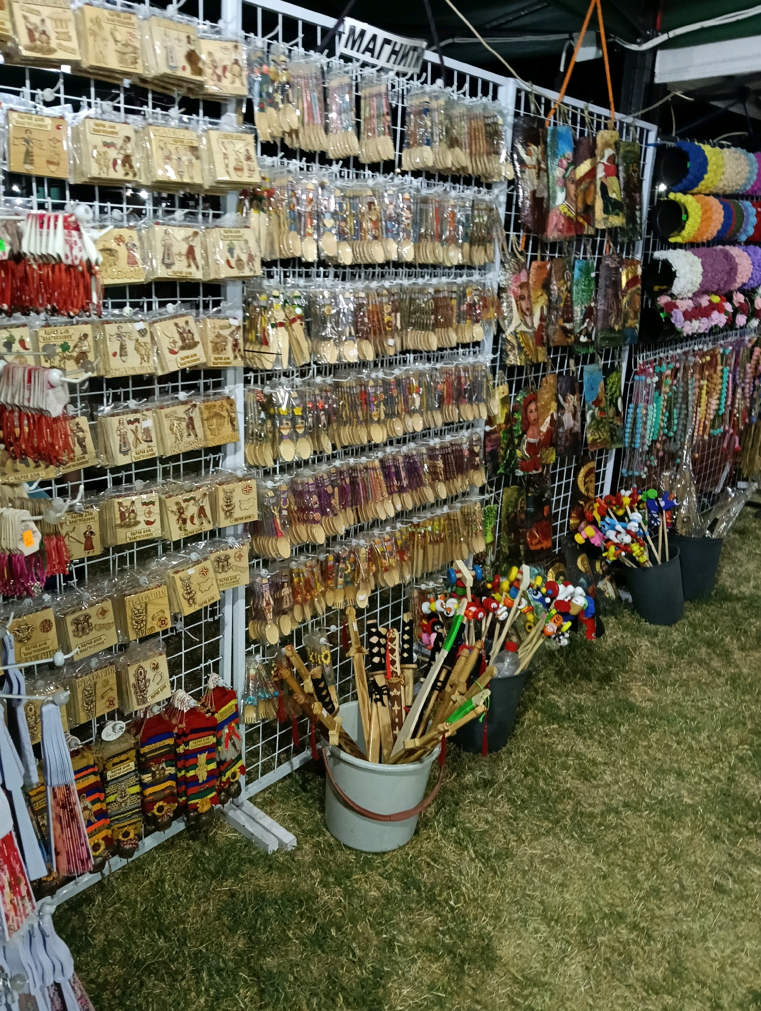 The image depicts a stall at an outdoor market, likely selling traditional crafts and souvenirs. The stall is organized with various items displayed on white wire racks. In the foreground, to the left, are folded paper fans. To the right of the fans, hanging from the racks, are colorful, woven textile items with bells attached. Further to the right, a bucket holds a collection of wooden items with painted figures on the ends, possibly toys or decorative pieces. Behind this, more buckets contain similar items, along with a plastic bottle. The stall is set up on a grassy area. The background shows more racks filled with a variety of goods, including small wooden plaques with designs, packets of wooden spoons and forks, beaded necklaces, and decorative hair accessories. The overall impression is one of a bustling market with a wide array of handmade goods.