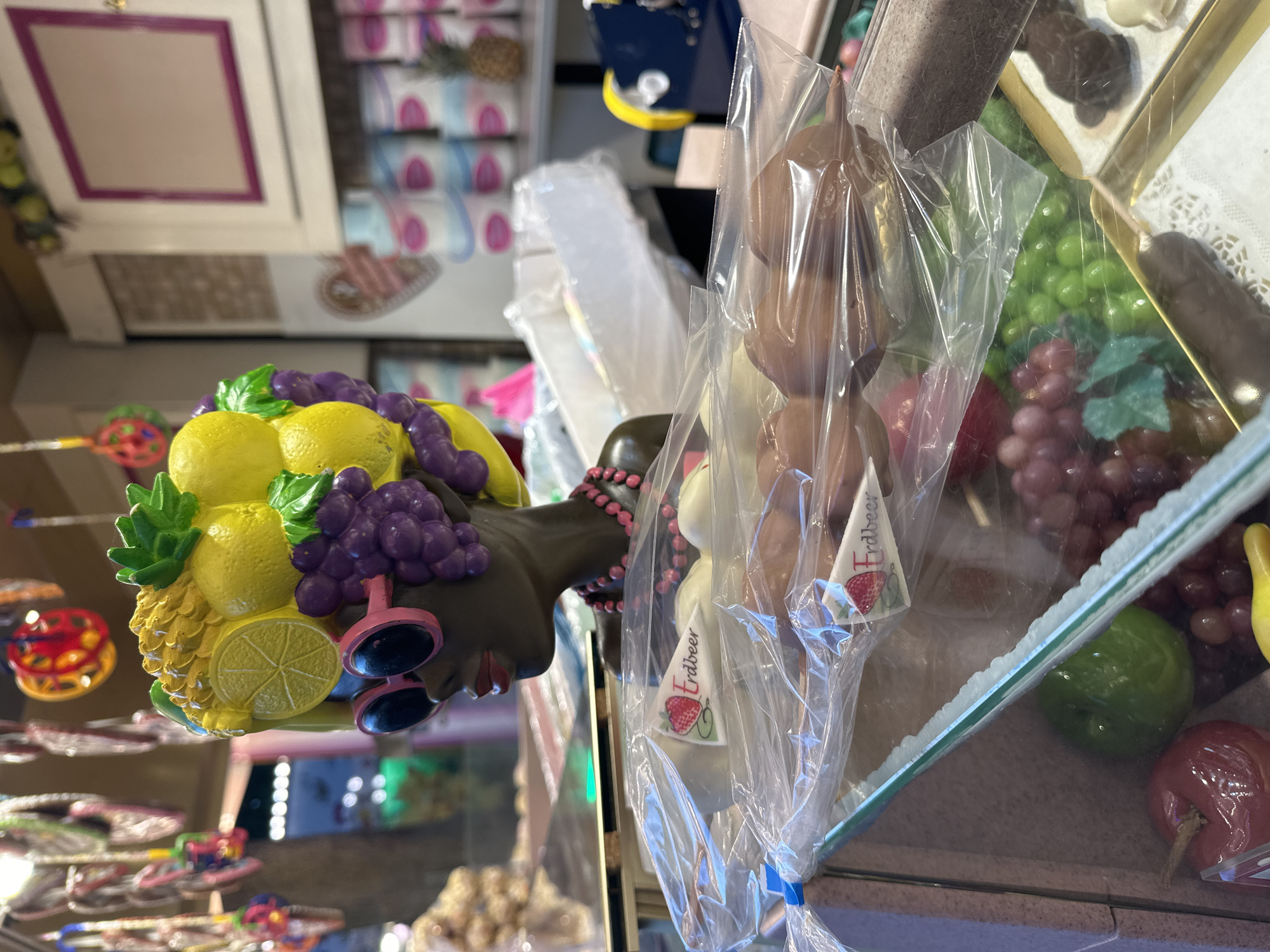 The image shows a display case at a confectionery shop in Bruchhausen-Vilsen, Germany. In the foreground, there are several individually wrapped chocolate-covered items on sticks, with labels that say "Erdbeer" (strawberry) and feature a strawberry illustration. Behind these, the display case contains various chocolate creations, including fruit shapes like apples and grapes, as well as chocolate figures. The backdrop reveals a counter with stacked goods, possibly candy or baked items, and decorative elements hanging from the ceiling. The overall scene suggests a festive and appealing presentation of sweets.