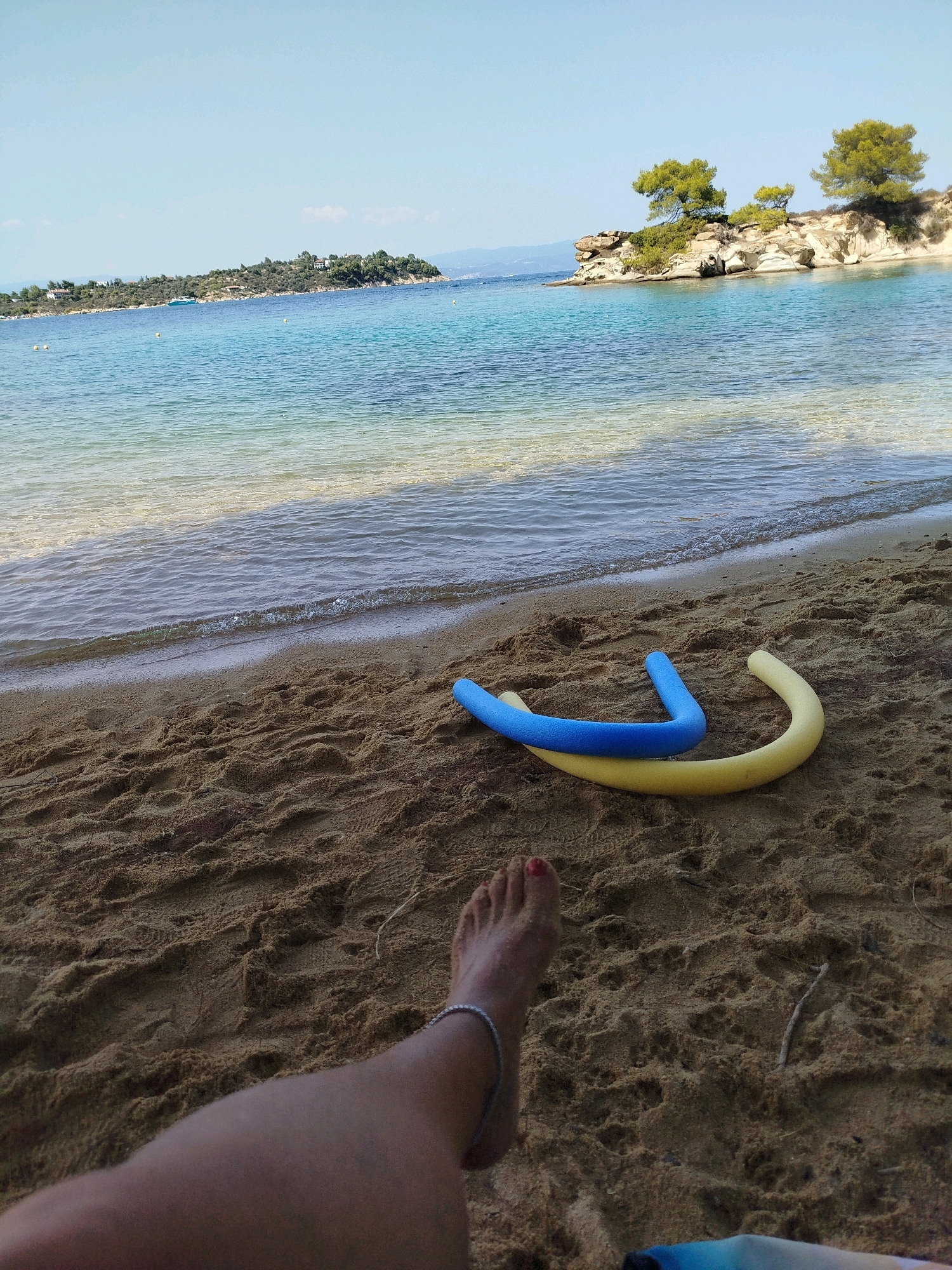 A person's foot, adorned with a silver anklet and red nail polish, rests on sandy ground near the edge of the turquoise waters of the Aegean Sea. Two pool noodles, one blue and one yellow, lie on the sand behind the foot. In the background, a small island covered in green vegetation and trees juts out into the sea, with more land visible in the distance under a clear blue sky. The scene captures a tranquil moment on a beach in Sithonia Municipal Unit, Greece.