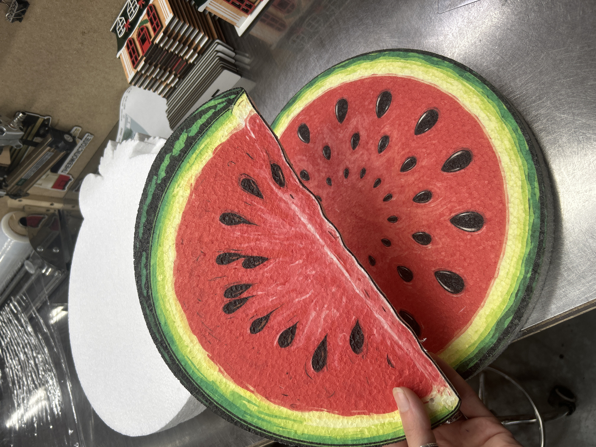 In Ternopil, Ukraine, a person's hand with a ring on their finger is holding a section of a large, circular, artificial watermelon slice. The watermelon slice is depicted with a bright red interior filled with black seeds, a yellow rind, and a green outer rind. Another, larger section of the same artificial watermelon slice rests on a metallic surface below. In the background, stacks of miniature houses are visible, along with some workshop equipment and rolls of plastic wrap. The overall scene appears to be in a workshop or production area.
