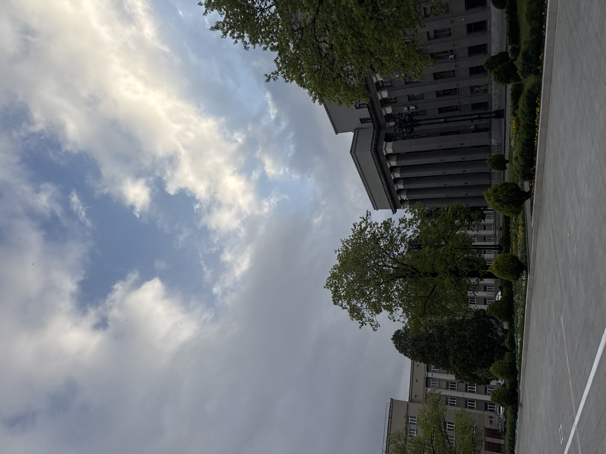 The image captures a view of a building in Akhieli, Georgia, situated under a cloudy sky. The building, constructed with grey materials, features classical architectural elements such as columns. Trees with green foliage are visible on either side of the building, with a paved area stretching out in the foreground. No people or explicit interactions are present. The environmental conditions suggest a possibly overcast day.