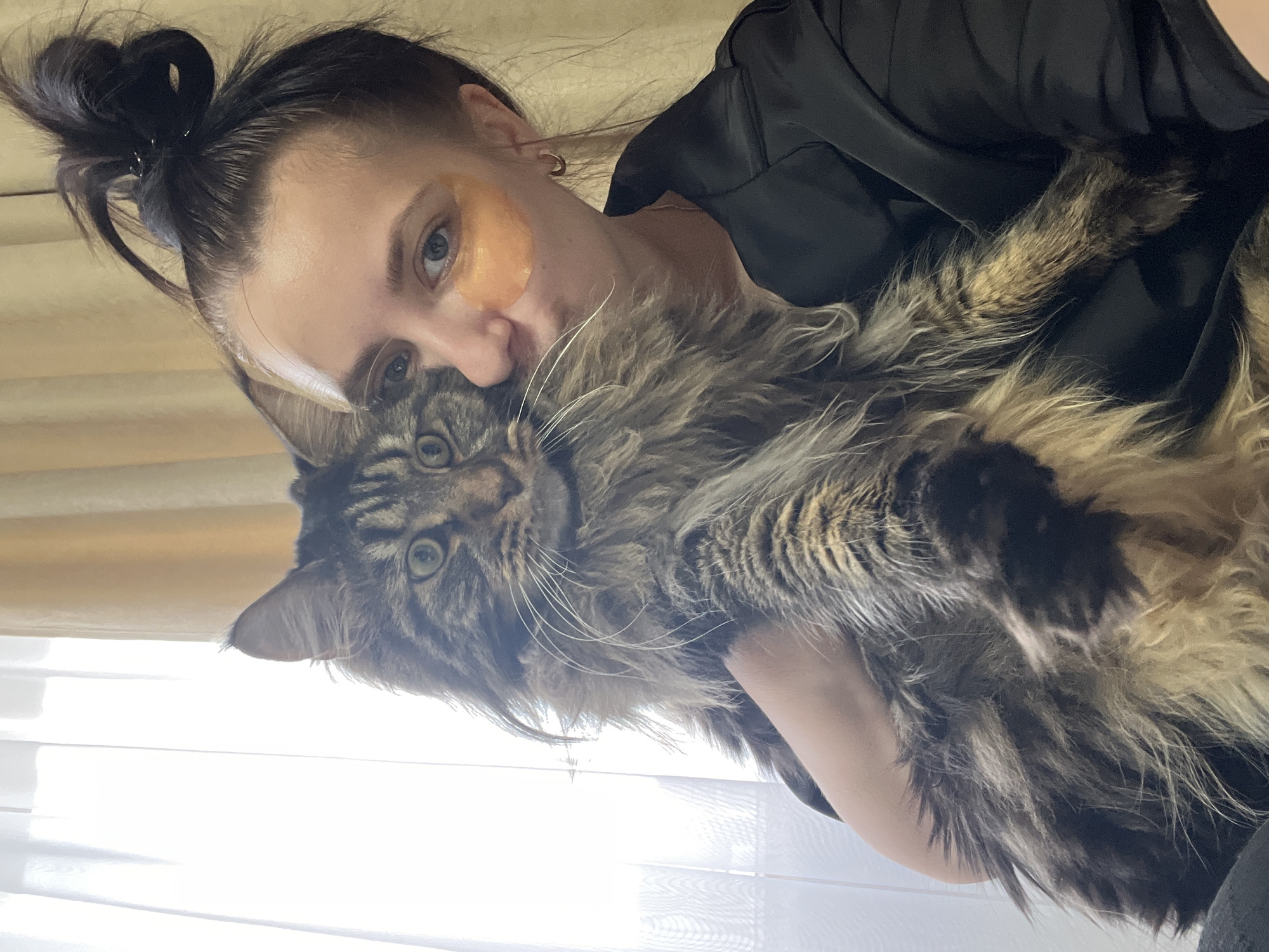 A woman with dark hair pulled into a messy bun and wearing under-eye masks holds a large, fluffy cat with tabby markings. The woman is leaning in to kiss the cat, who is looking towards the camera with wide, green eyes. She is wearing a black satin robe. The background shows a window with sheer white curtains and a beige curtain. The scene appears to be indoors, likely in a residential setting in Марковский, Russia.