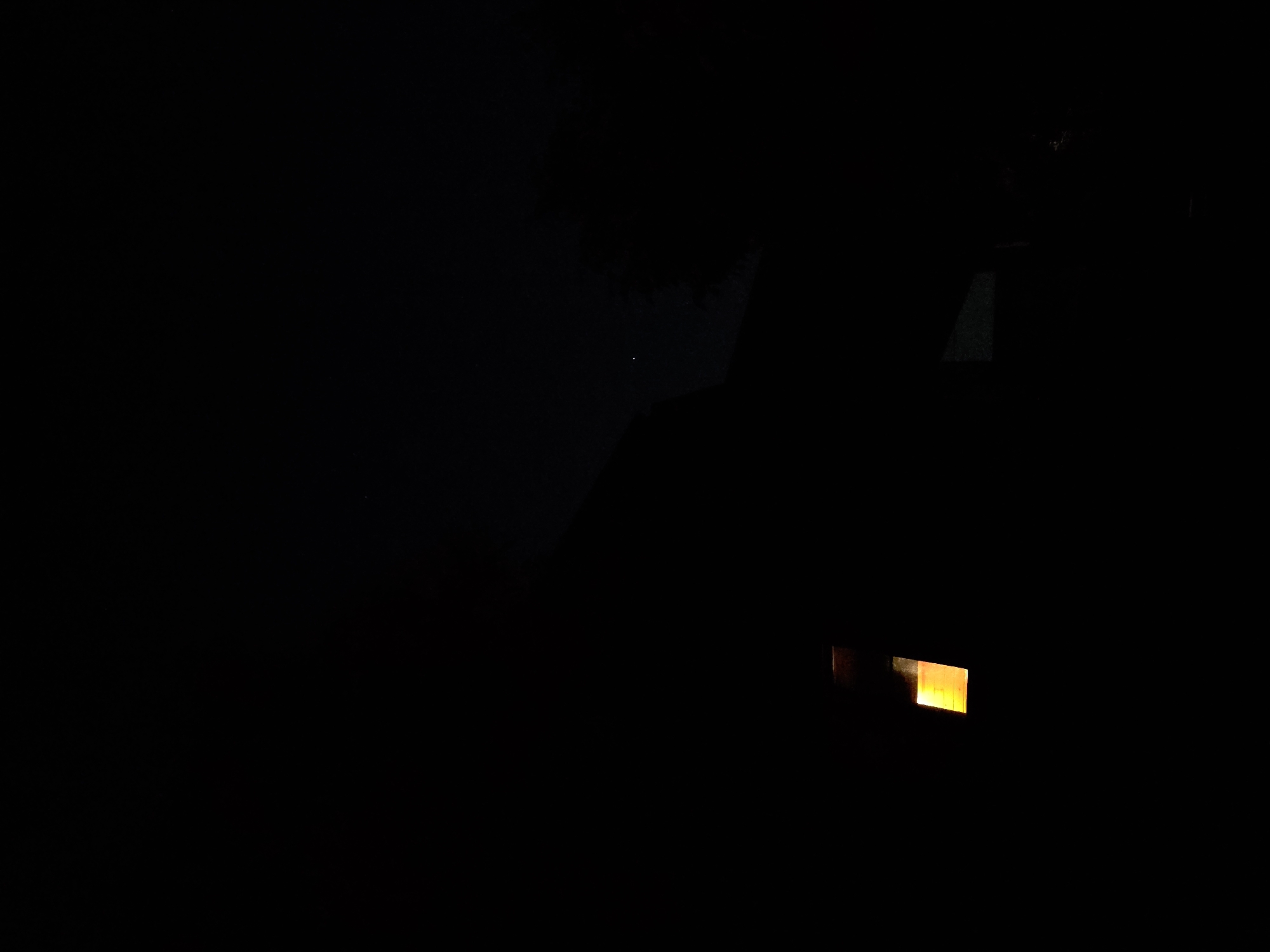 The image, taken at night in the area of Строитель-нефтяник Снт, Russia, depicts a dark, possibly rural or suburban landscape. The most prominent feature is a brightly lit rectangular object with an orange glow, positioned in the lower left quadrant. This object appears to be a light source, possibly a window or a lantern, casting a warm light against the surrounding darkness. In the upper right portion of the image, there is a faint, small white speck, which could be a distant light or a star. Silhouettes of what appear to be structures, possibly houses or trees, are vaguely discernible against the dark sky, suggesting the presence of buildings and natural elements within the scene. The overall atmosphere is one of nighttime tranquility, with the single light source serving as a focal point.