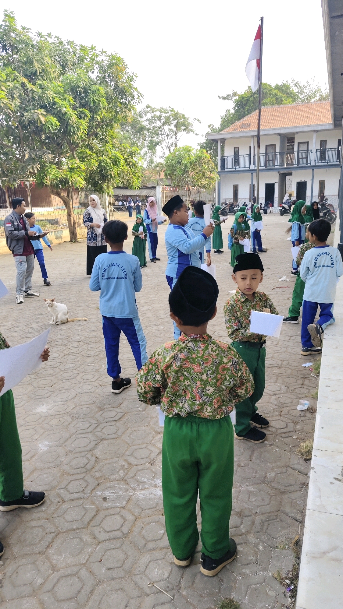 In Banyubunih, Indonesia, this image captures an outdoor scene, likely a schoolyard, with a group of children and adults. The children, dressed in school uniforms, appear to be participating in an outdoor activity. Some are wearing light blue shirts and blue pants, while others are in green outfits with patterned shirts. Several children are holding pieces of paper. One child in the foreground wears a black peci and a green patterned shirt with green pants. In the background, a white and red flag flies from a flagpole. A two-story building, possibly part of the school, is visible. A white cat is sitting on the paved ground. The environment is paved with hexagonal stones, and there are trees and fencing visible in the background. The overall atmosphere suggests an organized event or assembly.