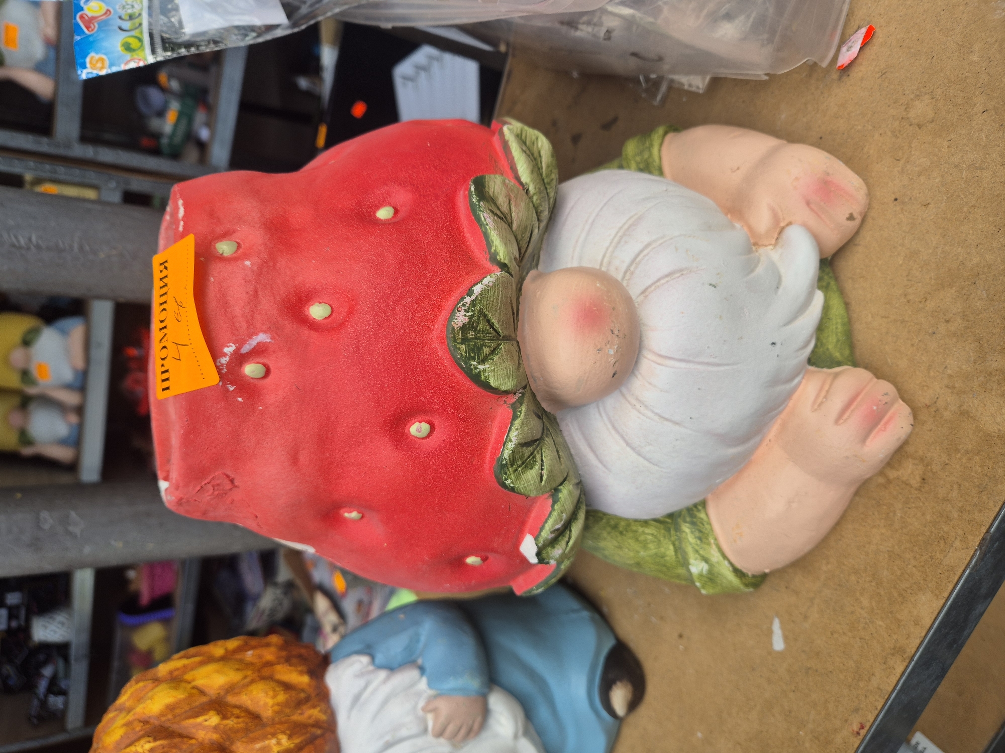 The image displays a close-up, top-down view of a garden gnome statue. The central subject is a gnome with a large, red strawberry serving as its hat. The strawberry features several light-green/yellow dots, resembling seeds, and green leaf-like structures where it meets the gnome's head. The gnome has a prominent white beard or hair, a light-pink nose, and bare skin-toned hands and feet. Its body appears to be clad in a green tunic.
The statue shows signs of wear and minor imperfections, such as scuffs on the nose, feet, and edges of the strawberry hat. An orange price tag is attached to the strawberry hat. The text on the tag, which is in Bulgarian, clearly reads "ПРОМОЦИЯ" (PROMOCIYA), meaning "PROMOTION." Below this, partially obscured numbers are visible, likely indicating a price such as "4.60" or similar.
The gnome is positioned on a light brown surface, possibly a shelf or display table, within what appears to be a retail environment. Metal shelving is visible in the background, along with blurred parts of other merchandise, including another gnome statue in blue and white, and a yellow/orange textured object, possibly another garden decoration. The overall scene suggests a display of garden ornaments or gift items, consistent with a store or market setting in Plovdiv, Bulgaria, as indicated by the Bulgarian text on the tag. No people are visible in the image.