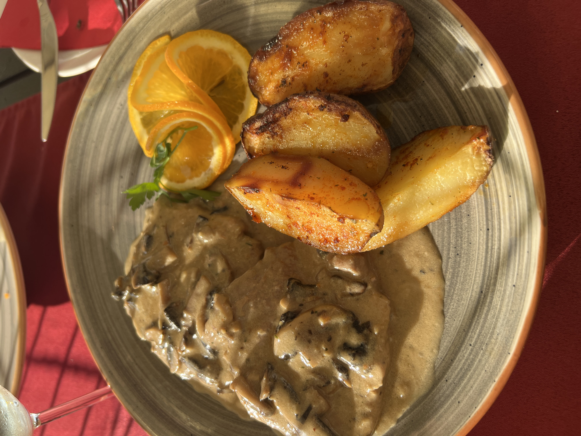 The image shows a close-up of a meal served on a plate. The dish consists of thinly sliced meat covered in a creamy mushroom sauce, accompanied by roasted potato wedges and garnished with orange slices and a sprig of parsley. The plate has a grey and white striped pattern with an orange rim and is set on a red tablecloth. The lighting suggests it is daytime, with sunlight casting shadows. The location is identified as Sveti Stefan, Montenegro.
