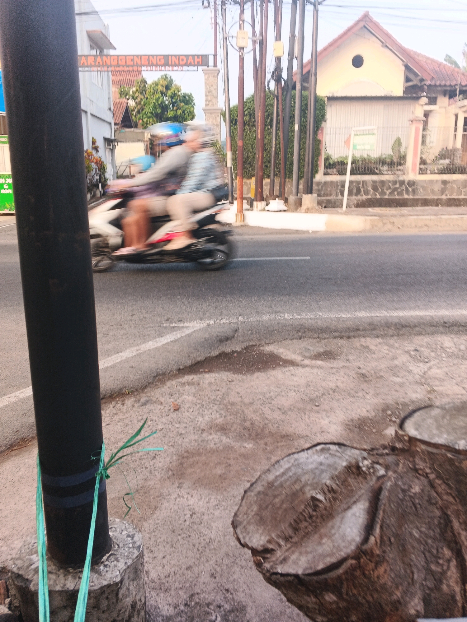 A blurred, medium-shot image taken at eye-level shows a street scene in Semarang, Indonesia. In the foreground, a black pole stands to the left of the frame, secured by green ties to a concrete base. To the right, the end of a tree stump is visible. The middle ground features a road with white lane markings, on which a motorcycle with two blurred figures is moving from left to right. The figures are wearing helmets. Behind the road, a building with a red tiled roof and a light-colored facade is visible, along with trees and electrical poles. An archway with the inscription "ARANGGENENG INDAH" is situated above the road. The overall scene suggests a street in a suburban or semi-rural area, possibly in the early morning or late afternoon due to the lighting.