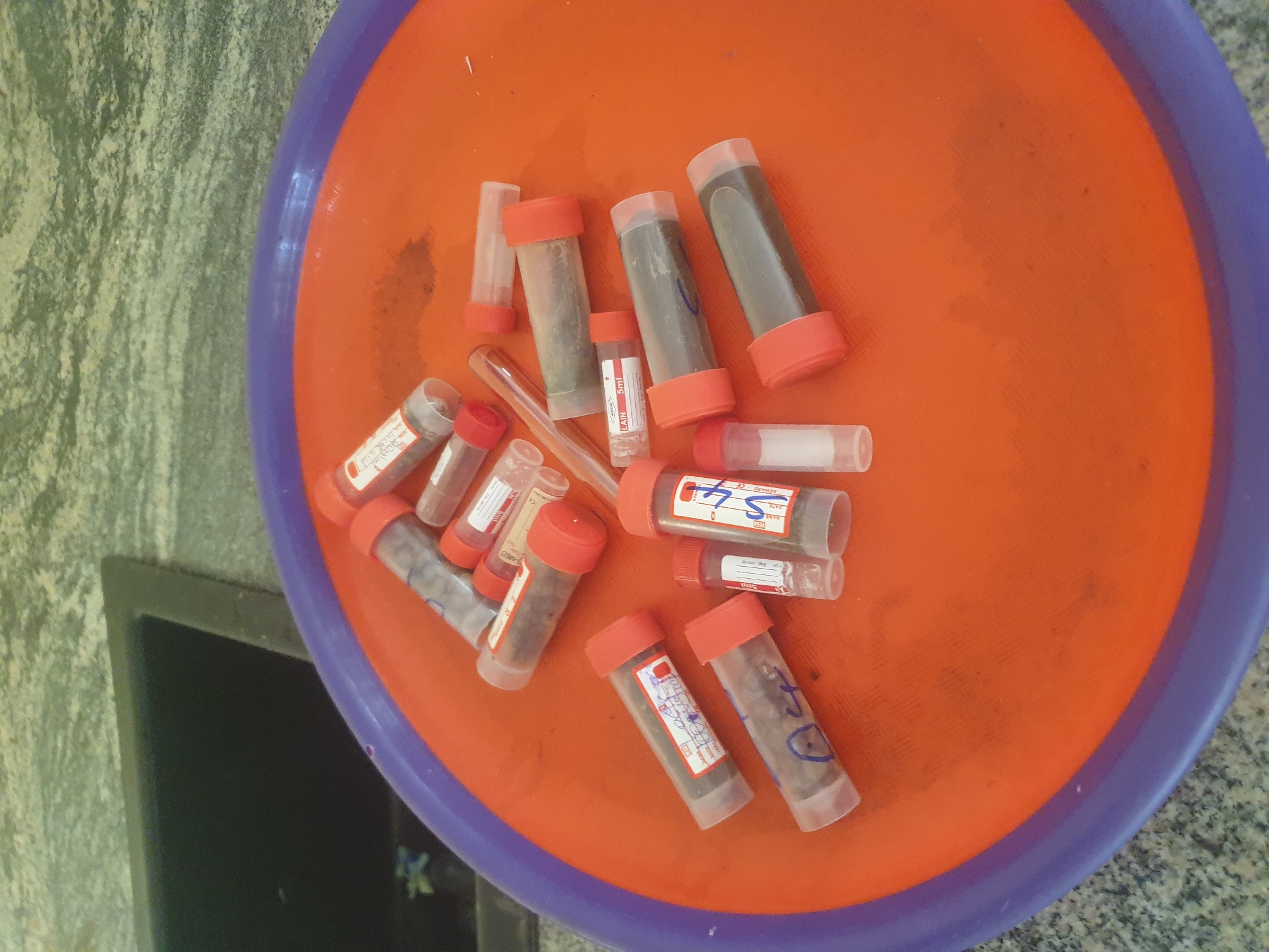 The image shows a collection of small, clear plastic vials with red caps scattered across an orange tray. The vials appear to contain various granular or powdered substances, some of which are dark brown or grey. Several vials have labels, some with handwritten text and dates. One vial is labeled "ST4" and has what appears to be a serial number. Another is labeled "LAIN 5ml". The tray is resting on a speckled, grey countertop. In the background, a black sink is visible. The scene is set in Jalingo, Nigeria.