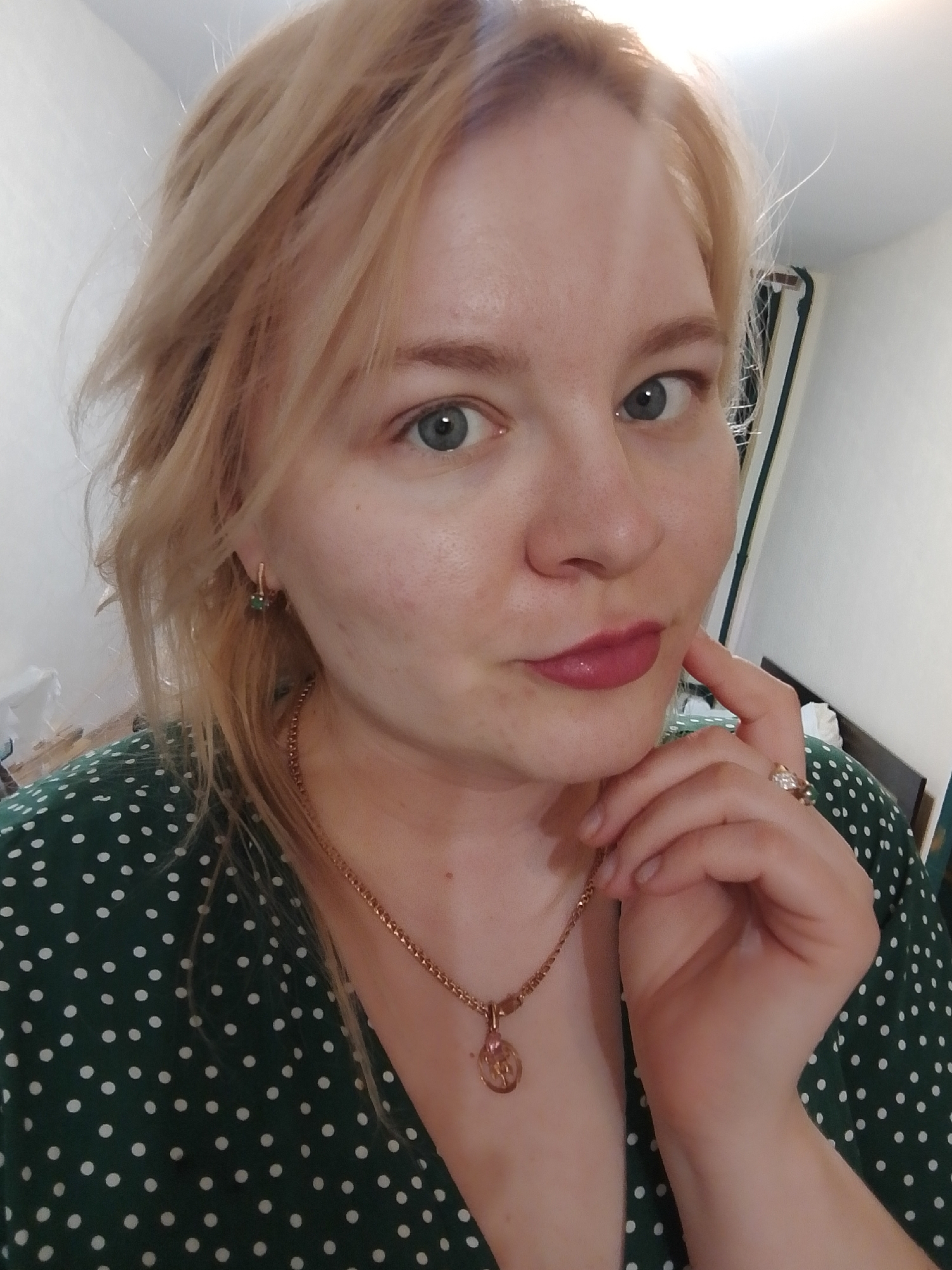 A fair-skinned woman with blonde hair and blue eyes is facing the camera. She is wearing a green dress with white polka dots, a gold necklace with a pendant, gold earrings with green stones, and a gold ring with diamonds. Her hand is positioned near her chin in a thoughtful pose. The background shows a white wall and a partial view of a curtained window. There is no indication of the location being Izhevsk, Russia, or any specific city.