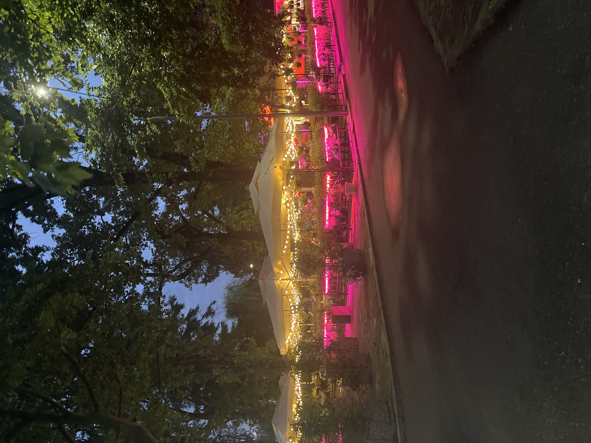 The image depicts an outdoor seating area of a restaurant or cafe in Krasnodar, Russia, at dusk. Several beige umbrellas are set up, providing shade over tables and chairs. String lights are draped around the umbrellas, creating a warm, festive atmosphere. The area is illuminated by pink and yellow lights, with the pink lights highlighting planters filled with greenery. The ground is paved with interlocking stones, and a street or pathway is visible in the foreground. Trees with lush green foliage surround the seating area, and the sky is a deep twilight blue. There are hints of people seated at the tables within the illuminated area, but they are not clearly visible. The overall impression is that of a pleasant evening setting for dining or socializing.