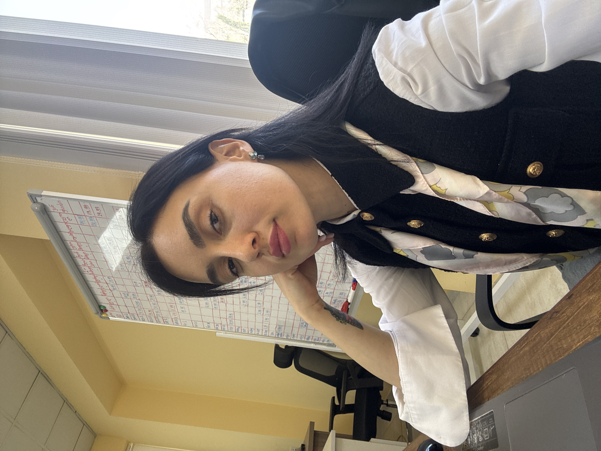 This media file presents a portrait of a young woman situated in what appears to be an office setting in Tbilisi, Georgia. The woman, with long dark hair, gazes directly at the camera, her head tilted slightly, and her left hand resting gently near her chin. Her expression is calm, with a subtle pout to her lips. She is dressed in a crisp white long-sleeved shirt layered under a black sleeveless vest adorned with multiple prominent gold-toned buttons. A light-colored scarf featuring a pastel, abstract pattern (possibly clouds or organic shapes) is tied around her neck. A small, light blue or green stud earring can be seen in her left ear, and a delicate butterfly tattoo is visible on her left forearm.
The background reveals typical office elements. To her left, a white dry-erase board is filled with handwritten information, including grids, numbers, and text. Visible numbers include "28.08 19.99," "29.08 19.99," and "28.09 19.99," among other entries in what appears to be a foreign language. The walls are a light yellow or cream color. Behind her, a window fitted with horizontal blinds suggests natural daylight is entering the room, indicating it is daytime. Parts of a dark office chair are visible behind her, and a dark brown wooden desk surface occupies the foreground to the right, with a corner of what might be a laptop or monitor. The overall lighting is bright and natural.