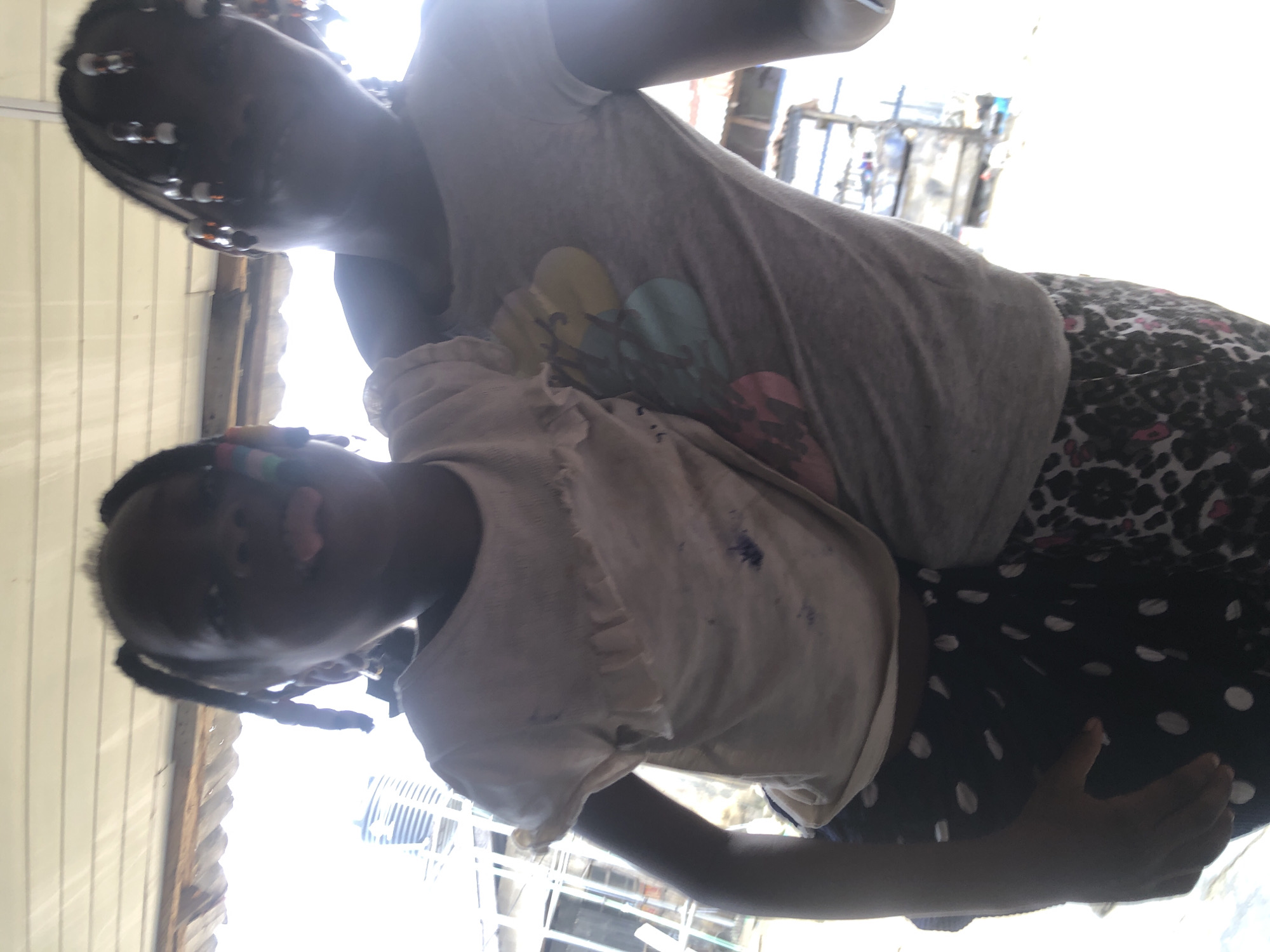 The image shows two young girls in an outdoor setting, likely in Ado, Nigeria. The girl in the foreground, on the left, is wearing a white t-shirt with ruffled sleeves and dark pants with white polka dots. She is holding the other girl and sticking her tongue out at the camera. The girl in the background, on the right, is wearing a grey t-shirt with colorful circular graphics and patterned leggings. She has her hair in braids adorned with beads. The background appears to be a somewhat cluttered outdoor area with corrugated metal roofing overhead and various structures, possibly market stalls or informal housing, in the distance. The lighting suggests it is daytime, with bright sunlight.