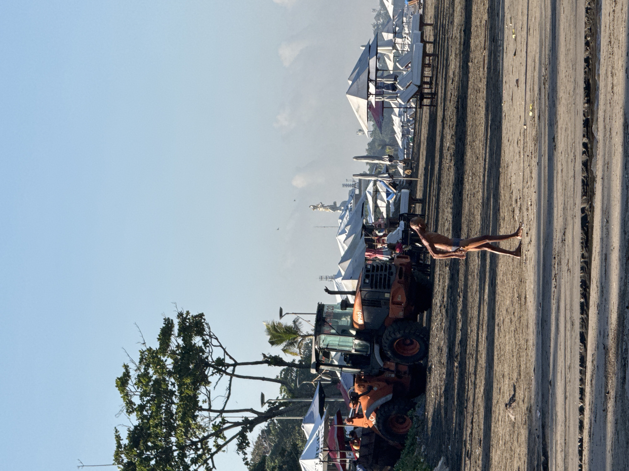 The image depicts a scene on Kuta Beach, Bali, Indonesia. A beach is seen with tire tracks in the sand. The tracks appear to be made by a large, orange construction vehicle that is parked by the beach. The vehicle is in the middle-ground to the left. A tree is also to the left, with its branches going towards the sky. A nearly nude person is seen lying down on the sand. In the background, there are a lot of white umbrellas and benches, some of which are set up under tents. In the distant background is a light, hazy horizon. The sky is light blue and mostly clear.