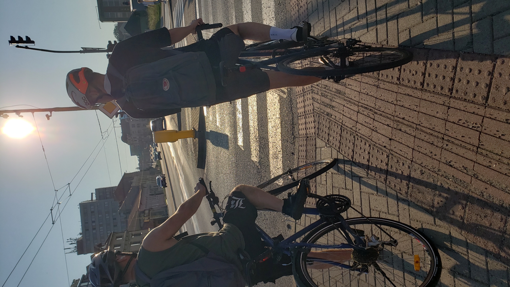 The image shows two individuals on bicycles at a pedestrian crossing in the city of Prešov, Slovakia. The sun is low in the sky, casting long shadows on the brick pavement and the street. Both cyclists are wearing helmets and backpacks. The person on the left is wearing a green tank top and black shorts. The person on the right is wearing a black t-shirt and dark shorts, with a distinctive white and orange helmet. In the background, buildings and a traffic light are visible, suggesting an urban environment. The road has several cars and what appears to be overhead electrical lines for public transport. The pedestrian crossing is marked with white stripes.