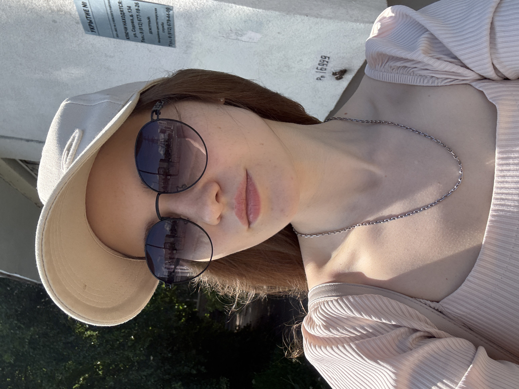 A young woman is captured in a close-up, outdoors, on a bright sunny day in Saint Petersburg, Russia. She wears a light beige baseball cap, dark round sunglasses, a light pink ribbed top, and a thin silver chain necklace. Her hair is light brown and flows over her shoulders. The photo is taken from a slightly low angle, with her head tilted, suggesting she is posing.
The background features a light-colored building wall, with some green foliage visible at the bottom and left side, indicating trees or bushes. Bright sunlight casts subtle shadows, highlighting the textures of her clothing and the wall.
Visible text on the wall in Russian includes information about a shelter: "УКРЫТИЕ №" (Shelter No.), along with details on where keys are located: "КЛЮЧИ НАХОДЯТСЯ: ул. Седова, д. 134" (Keys are located: Sedov St., 134). Contact numbers for a duty officer and emergency dispatch service are also listed: "Тел.: 8 (812) 677-18-26", "ДЕЖУРНЫЙ" (Duty officer), "Аварийно диспетчерская служба" (Emergency dispatch service), and "Тел.: 8 (812) 679-44-00". The area code "812" confirms the Saint Petersburg location. Another marking "P. 16999" is also visible on the wall.