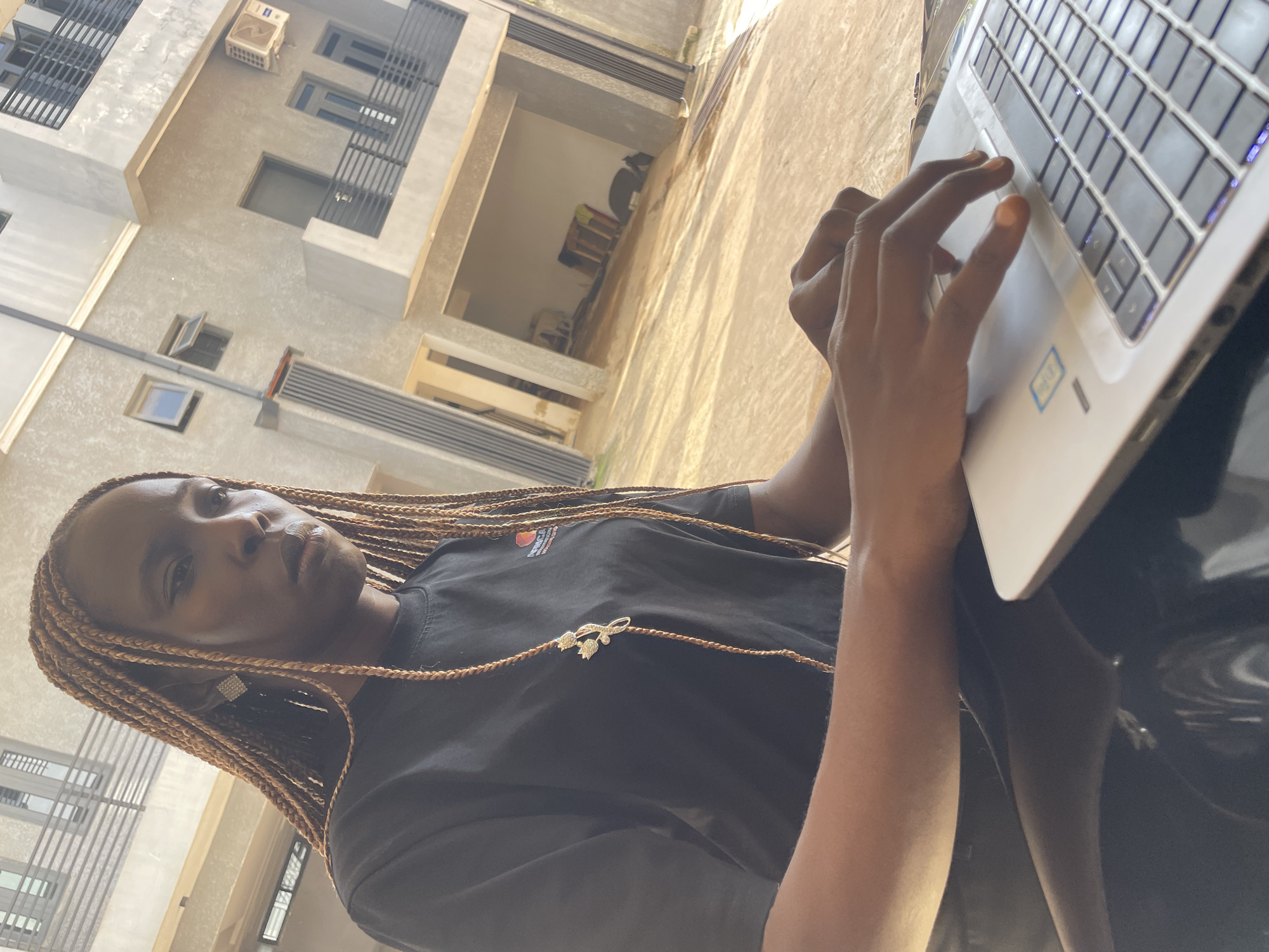 A medium shot shows a young Black woman with braided hair, wearing a black t-shirt, sitting outdoors and typing on a silver laptop. The woman is positioned in the left half of the frame, her head tilted down and eyes focused on the laptop. She has prominent cheekbones and a serious expression. Her hair is styled in long, thin braids that extend past her shoulders, with a decorative gold hairpin attached to one braid. She is wearing a square, diamond-encrusted earring in her left ear. The laptop is open and positioned on a dark surface in the foreground. Sunlight casts shadows across the scene.
In the background, a modern, multi-story building is visible. The building has a textured gray facade, with balconies featuring black metal railings. Several windows and doors are present, and an air conditioning unit is mounted on one of the walls. The environment appears to be an outdoor urban setting, possibly a residential or commercial area in Abuja, Nigeria, given the architectural style and the sunlight. The ground is paved with light-colored material, and there are some indistinct objects and shadows in the background, suggesting an open area like a courtyard or parking space.