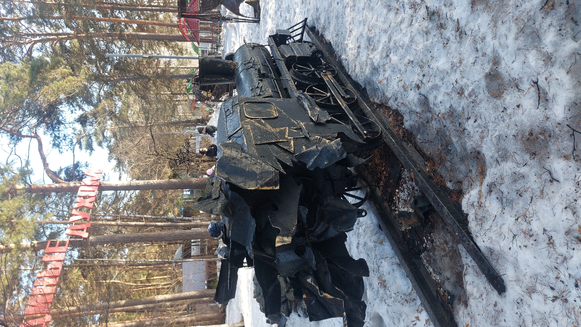 A full shot, at a slight downward angle, features a large, black metal sculpture of a steam train on a snow-covered ground, with trees in the background. The sculpture has a roughly textured surface and appears somewhat abstract, with sharp edges and angular shapes. The train is situated on metal tracks that are partially buried in the snow.
The background shows a forest with tall, slender trees, some of which have bare branches. A red sign is strung between the trees. People are visible in the background, their features indistinct due to the distance. The scene is set in a park in Novosibirsk, Russia. The ground is covered in patchy snow, suggesting a cold, potentially winter-like environment.