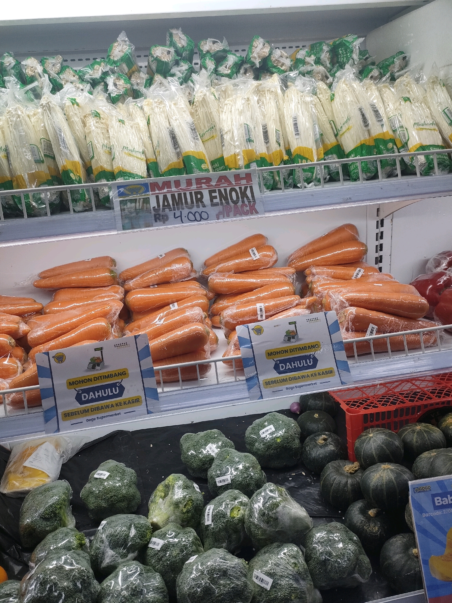 The image displays a grocery store refrigerated display case in Batam, Indonesia, showcasing various fresh produce. In the top section, rows of packaged enoki mushrooms are visible, with a prominent sign displaying "MURAH JAMUR ENOKI RP 4.000 PACK." Below this, a large quantity of fresh carrots are neatly arranged and individually wrapped in plastic. A blue and yellow sign is placed in front of the carrots, reading "MOHON DITIMBANG DAHULU SEBELUM DIBAWA KE KASIR," which translates to "Please weigh first before bringing to the cashier," followed by "Gogo Supermarket." The bottom section of the display features packaged broccoli florets and several dark green pumpkins or squash. A red plastic basket is also visible on the right side of the frame. The environment appears to be a well-lit supermarket aisle.