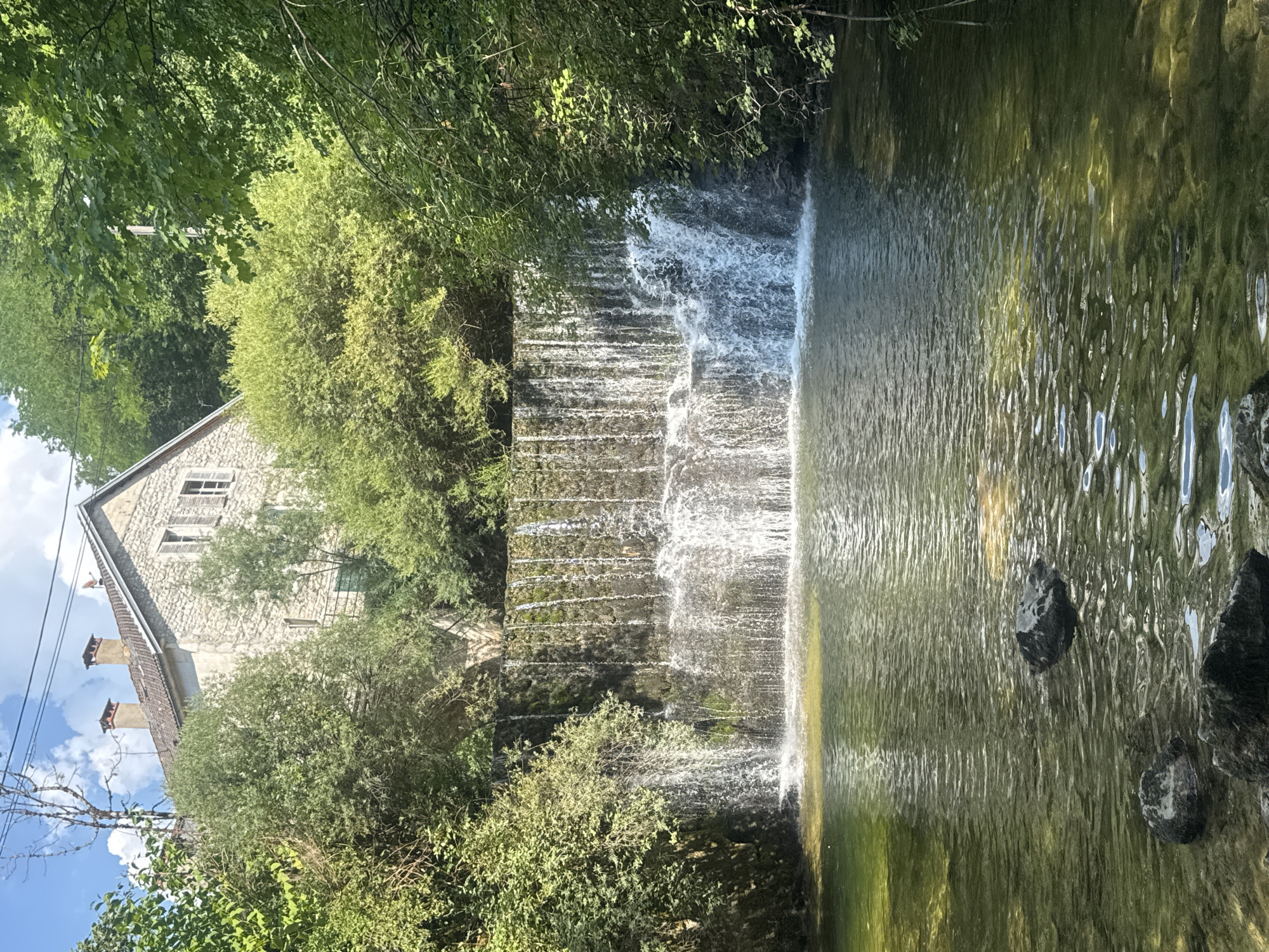 The image depicts a tiered waterfall flowing into a calm body of water, with lush green foliage surrounding the scene. To the left, a stone building with a red-tiled roof and multiple chimneys is visible, nestled amongst the trees. The building appears to be old, with some windows boarded up. Power lines are also visible running across the left side of the frame, partially obscuring the sky.
The waterfall itself is a prominent feature, with water cascading down a series of rocky steps, creating a dynamic visual effect. The water in the pool appears greenish and reflects the surrounding greenery and sunlight. Several dark rocks are scattered within the pool near the waterfall's edge.
The overall impression is one of a natural, serene setting, perhaps in a rural or semi-rural location. The abundance of trees suggests a temperate climate, and the bright sunlight indicates it is daytime. The image evokes a sense of peace and tranquility. The location is identified as Saint-Claude, France. There is no visible text within the image.