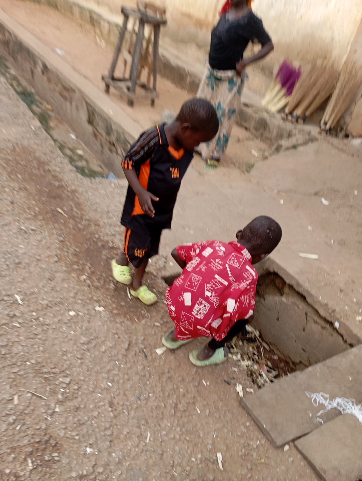In Shendam, Nigeria, two young boys are the focal point of this image, captured in an outdoor setting. The boy on the left, dressed in a black and orange t-shirt and shorts, stands with his head bowed, seemingly interacting with the other child. He wears bright yellow slip-on shoes. The boy on the right is crouched down, looking into a drainage ditch. He is wearing a vibrant red shirt with a white patterned design and green slip-on shoes. The environment appears to be a dusty, unpaved area, with a concrete drainage channel running alongside. In the background, a woman in a black top and a patterned skirt stands near a collection of folded items, possibly umbrellas, and a wooden structure that resembles a crude hoist or scaffolding. The overall impression is a candid snapshot of everyday life in a Nigerian town.