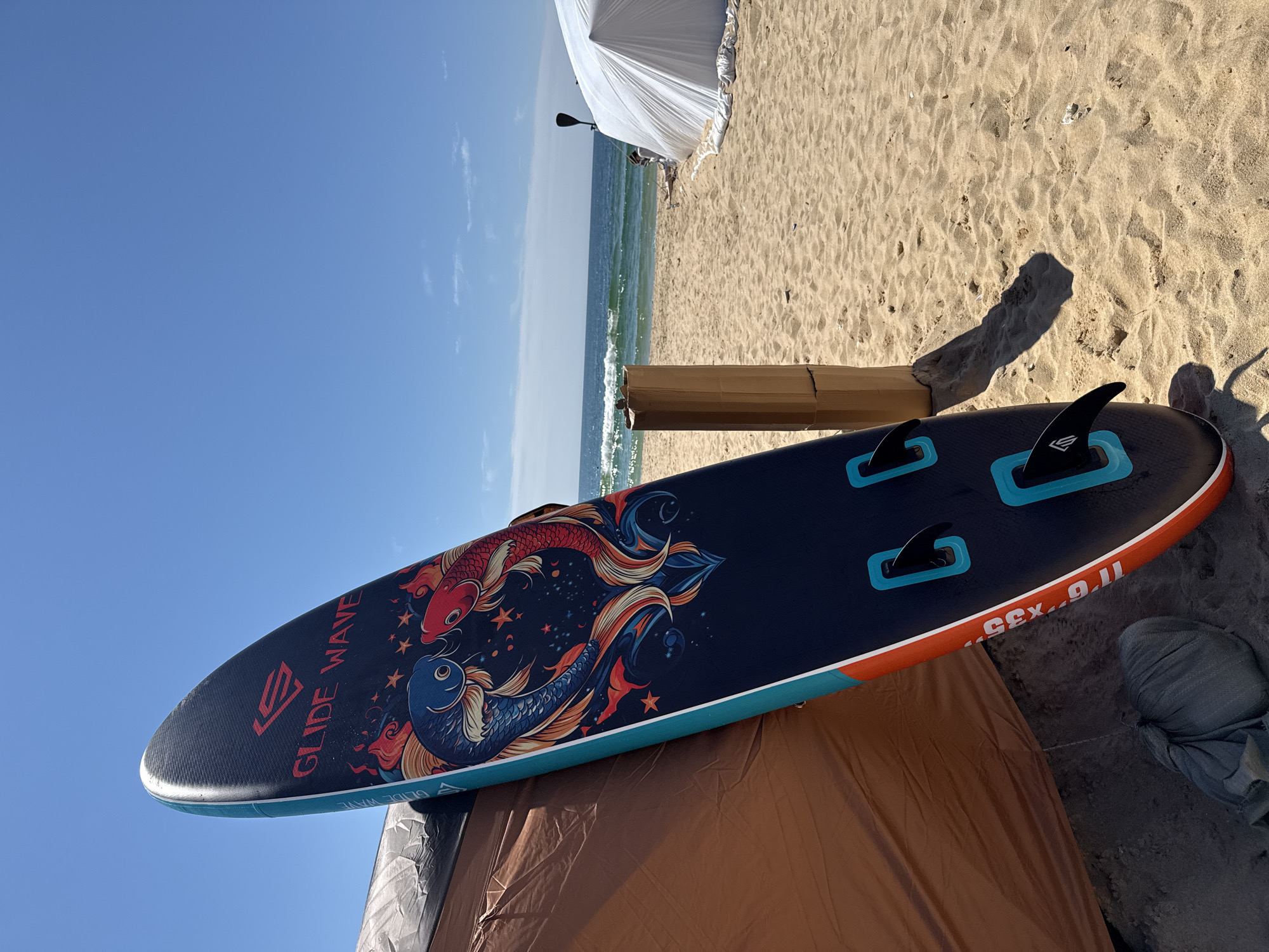 An inflatable paddleboard with a koi fish design stands upright on a sandy beach. The paddleboard is leaning against a tan tent. In the background, the ocean meets the horizon under a clear blue sky. Another white tent is visible further down the beach, along with a paddleboard oar. The sand is light brown and textured with ripples.