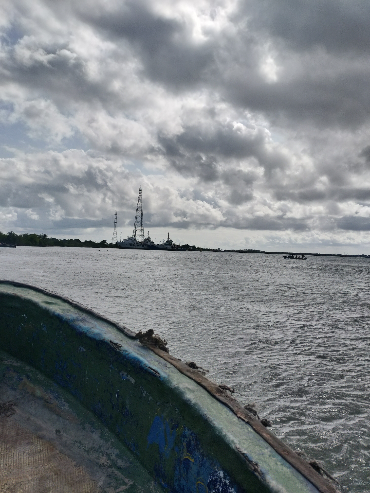 The image depicts a view of the Lagos Lagoon in Lagos, Nigeria, from the perspective of a boat. The foreground shows the edge of a weathered, green and blue painted boat. In the mid-ground is the open water of the lagoon. In the distance, the skyline includes vegetation, industrial structures including communication towers and cranes, and docked vessels. A small boat carrying several people is visible in the water. The sky is overcast with dark, heavy clouds.