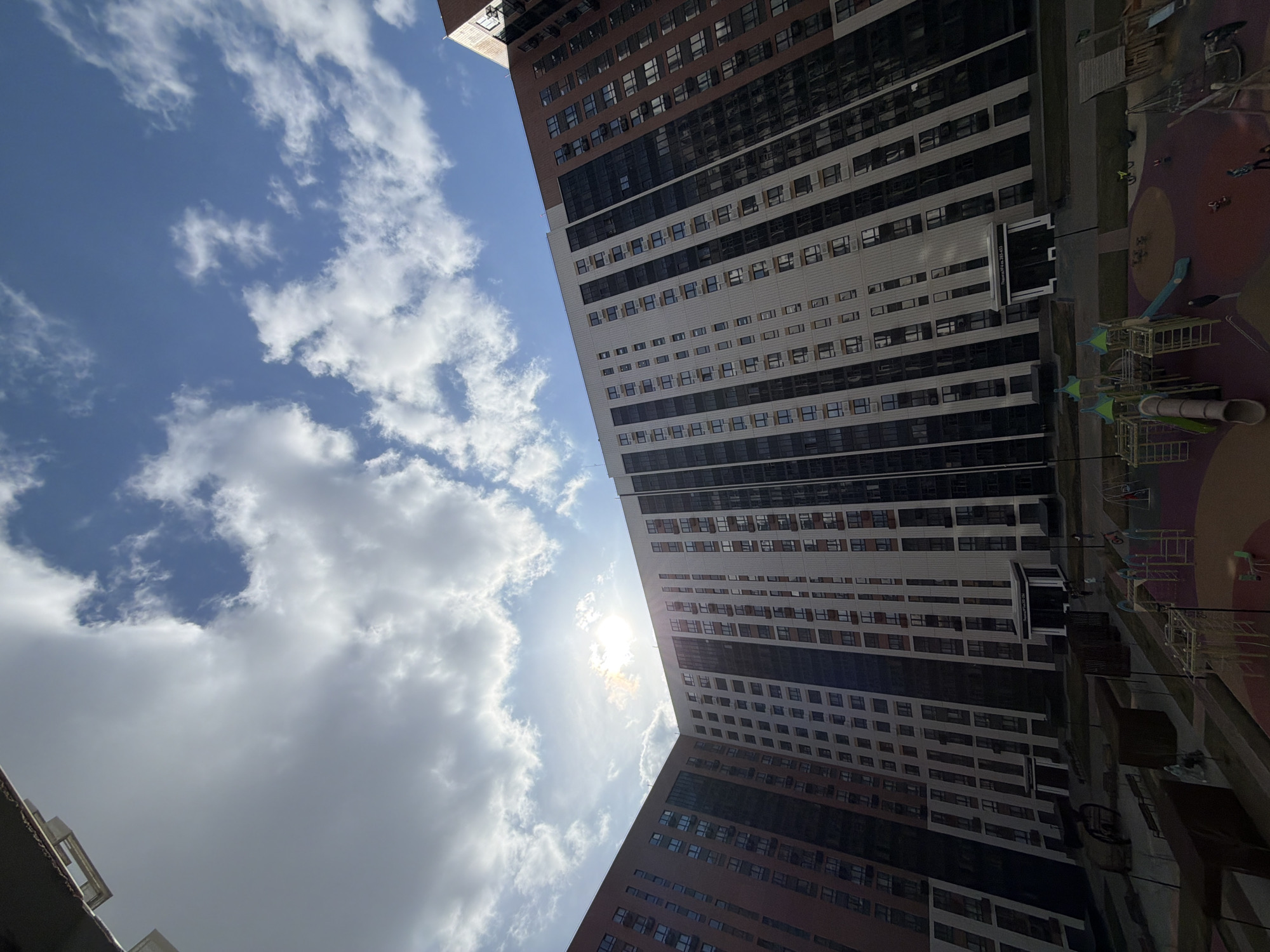 The image shows a low-angle view of tall buildings in Voronezh, Russia. The sky is partly cloudy with the sun visible. To the right of the buildings is a playground.