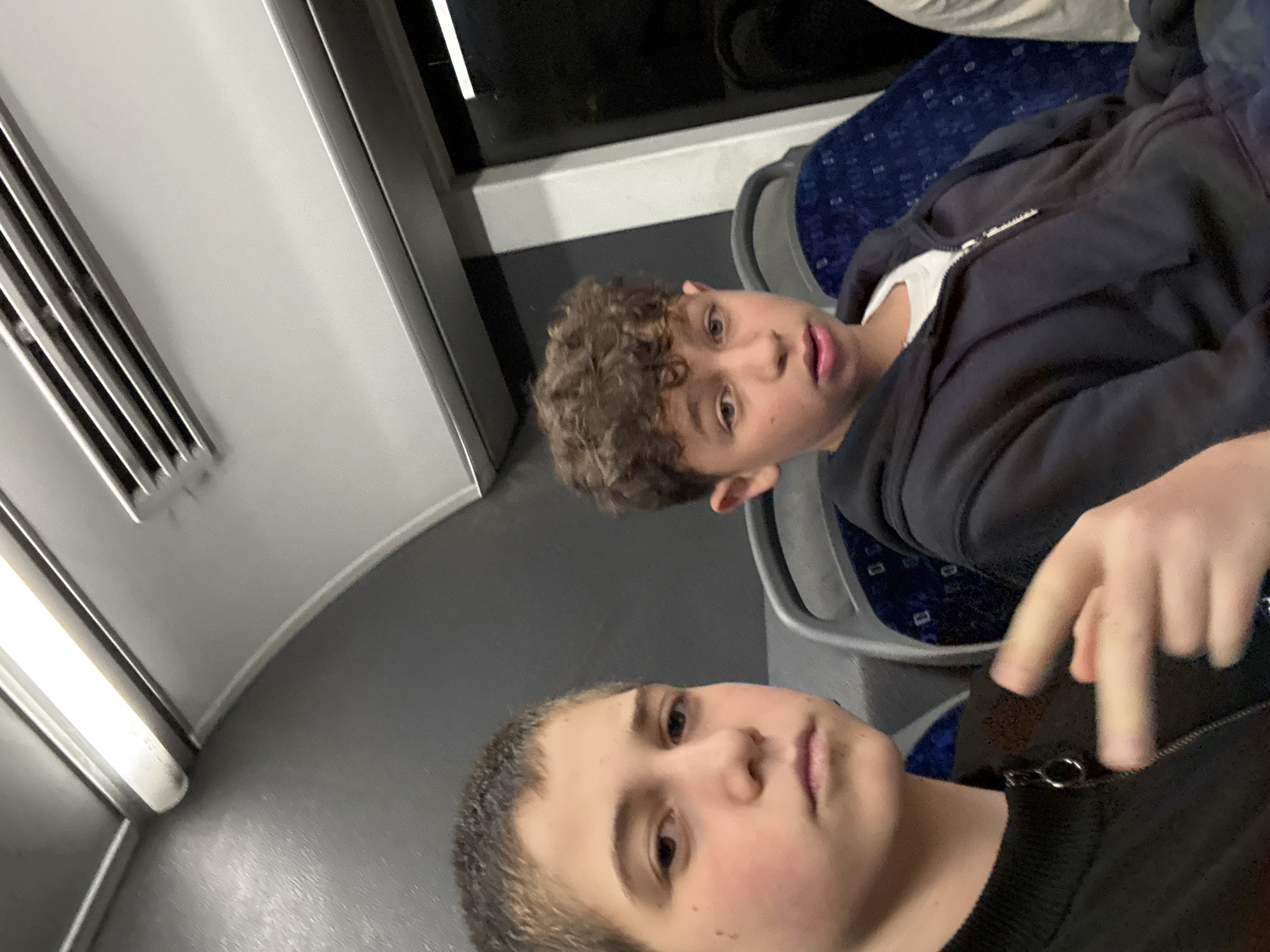 Two boys are on a train. One has curly brown hair and is wearing a black zip-up hoodie over a white t-shirt. The other boy has short brown hair and is wearing a black sweater and a black zip-up hoodie. They appear to be taking a selfie. The train car is gray and white with blue patterned seats. It is not possible to determine if the location is Tel-Aviv or any specific place in Israel from the image.