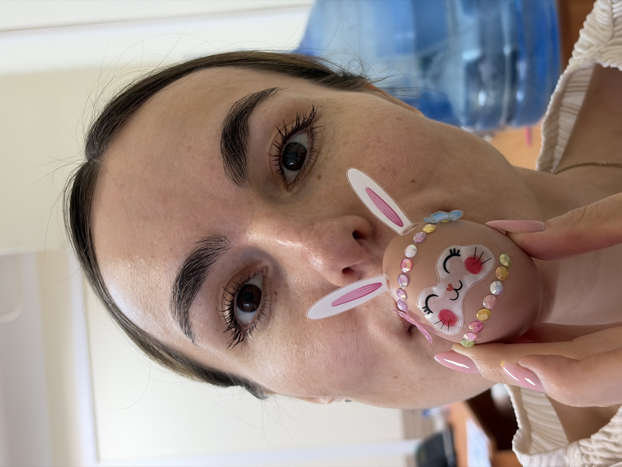 A woman is holding an Easter egg up to her face. The egg is decorated with a rabbit face, ears, and colored jewels. The woman has dark hair and brown eyes. She is wearing a white top. The background is out of focus, but appears to be an indoor setting with a large water cooler in the background. No information is available to suggest the location is Izhevsk, Russia.
