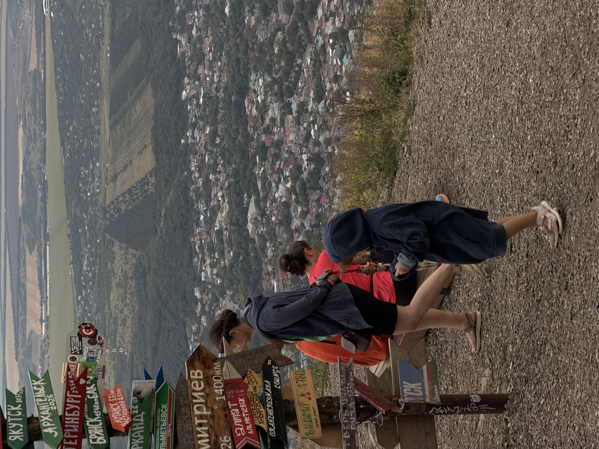 The image shows a panoramic view of a city nestled in a valley, likely Goryachevodsky, Russia, from a high vantage point. In the foreground, a weathered wooden signpost is adorned with numerous directional signs pointing to various locations, some with distances indicated. Three individuals are visible, walking or standing near the signpost. A woman in a dark blue hoodie and black shorts is in the center, looking away from the camera. Behind her, a person in a bright pink shirt is also present. A child in a dark blue hooded sweatshirt is to the right, facing away from the viewer. The ground is covered in gravel and dry grass. The city below is characterized by a dense arrangement of houses with red and gray roofs, interspersed with greenery and trees. The landscape suggests a mountainous or hilly terrain surrounding the urban area.