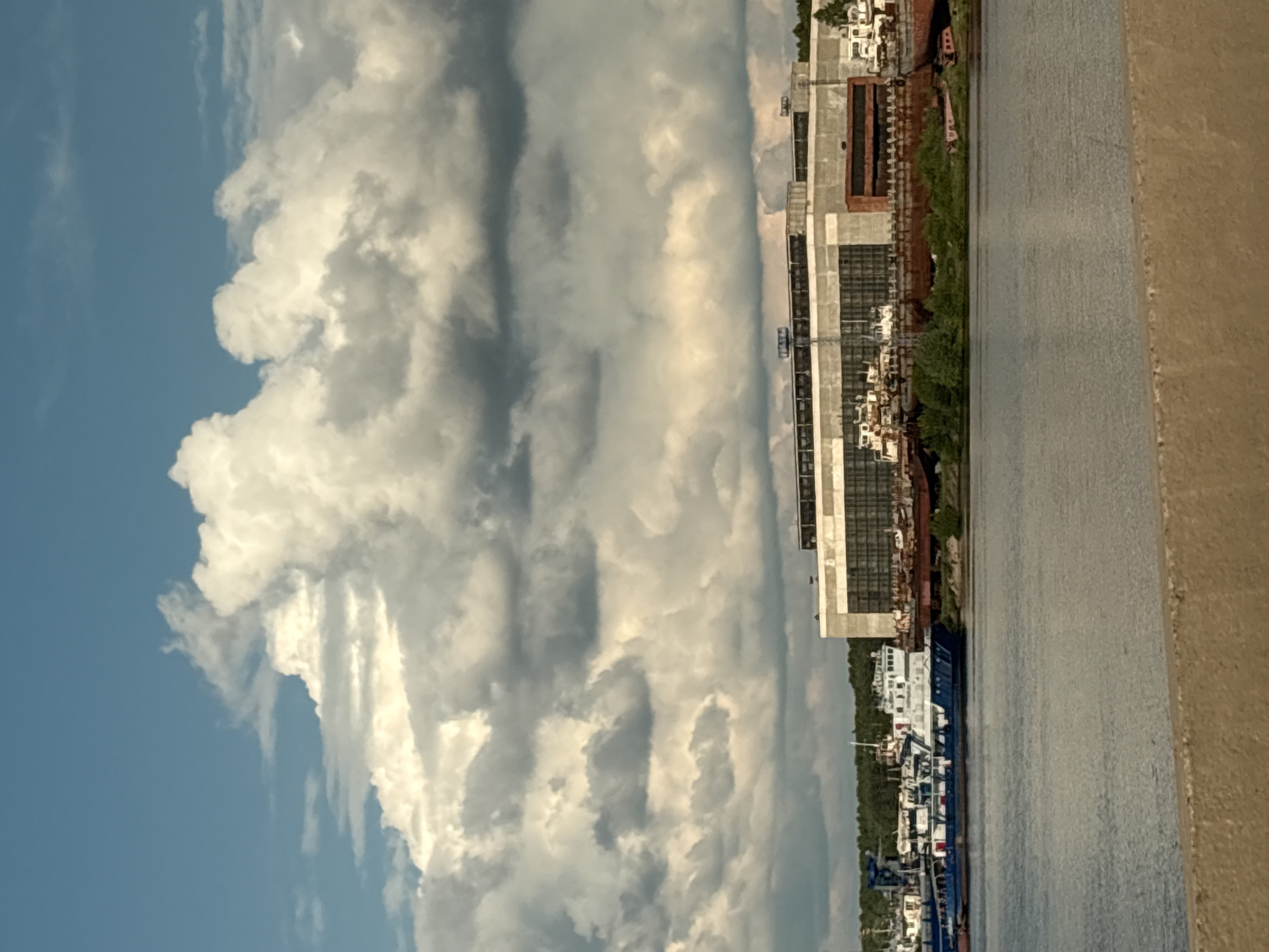 The image captures a wide-angle view of a river or canal waterfront in Chaikovsky, Russia, under a partly cloudy sky. In the foreground, a broad, light-brown/beige concrete or dirt embankment stretches across the bottom right, suggesting a viewing platform or a bank.
The midground features a wide expanse of calm, grey-blue water, reflecting the sky above. On the opposite bank (left side of the image), a landscape of industrial or commercial buildings, some appearing to be under construction or derelict, lines the shore. These structures are made of concrete and metal, with visible scaffolding and various components. Green trees and vegetation are scattered among the buildings and along the waterline.
Prominently featured on the water, near the center-left, is a large, multi-decked blue and white ship or barge, possibly docked or slowly moving. Other smaller vessels or structures can be seen further along the bank.
The background is dominated by a vast, bright blue sky filled with impressive, voluminous white and grey cumulus clouds that stretch across the horizon. The lighting suggests it is daytime, with clear visibility and distinct shadows indicating a strong light source. No people or visible text are discernible in the scene. The overall impression is one of an active, industrial waterway under a dynamic sky.