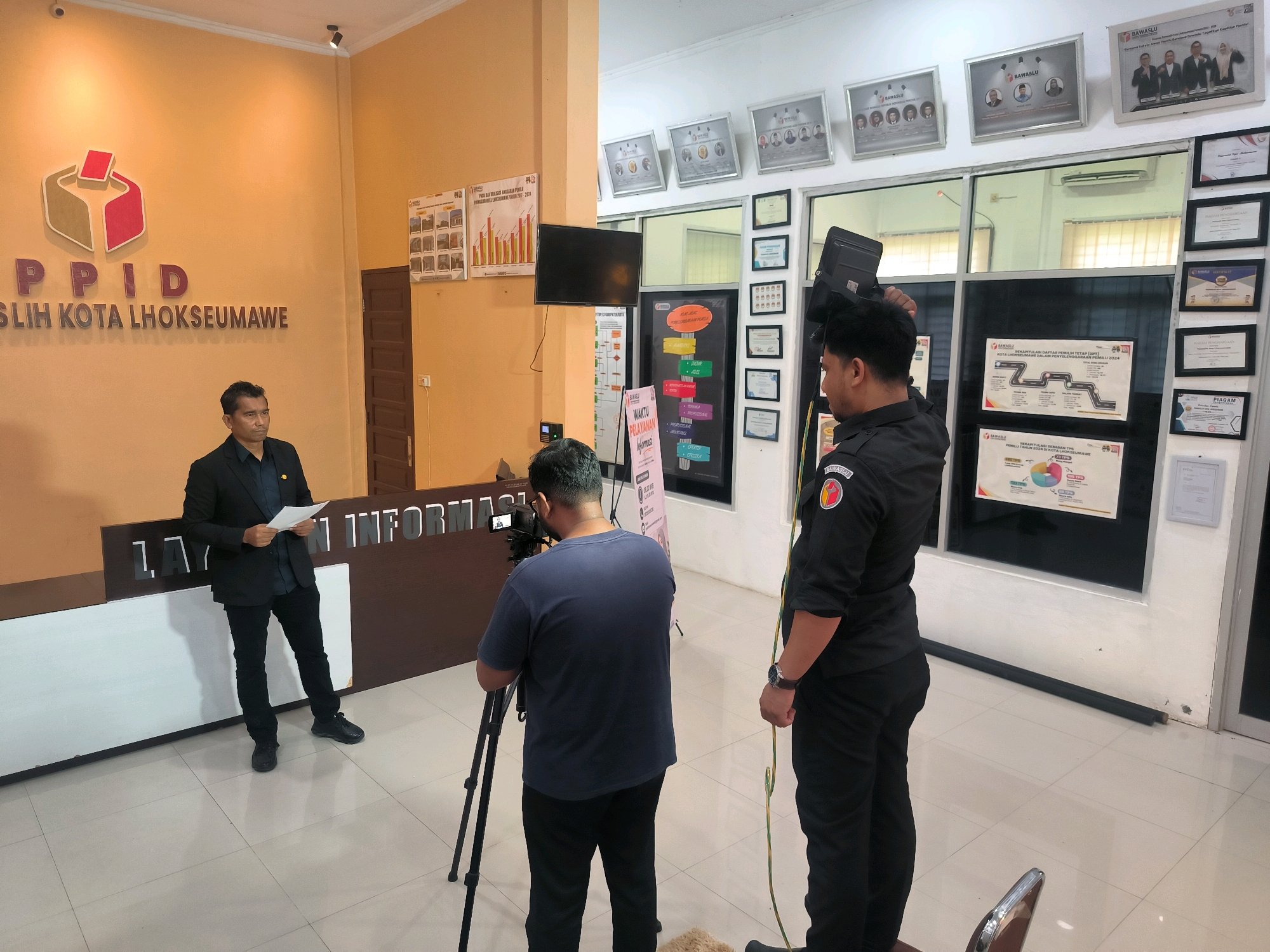 The image depicts an indoor scene, likely within an official building in Lhokseumawe, Indonesia. On the left, a man in a dark suit stands behind a reception desk, holding a document. The desk has the text "LAYANAN INFORMASI" visible. To the right of the reception desk, a man in a dark uniform, possibly security or event staff, holds a black spotlight. In the foreground, a tripod with a camera is set up, and a person wearing a dark blue t-shirt is operating it. The walls are painted orange and white, and several framed certificates and charts are displayed. The overall impression is of an organized event or informational session taking place within a public service area.